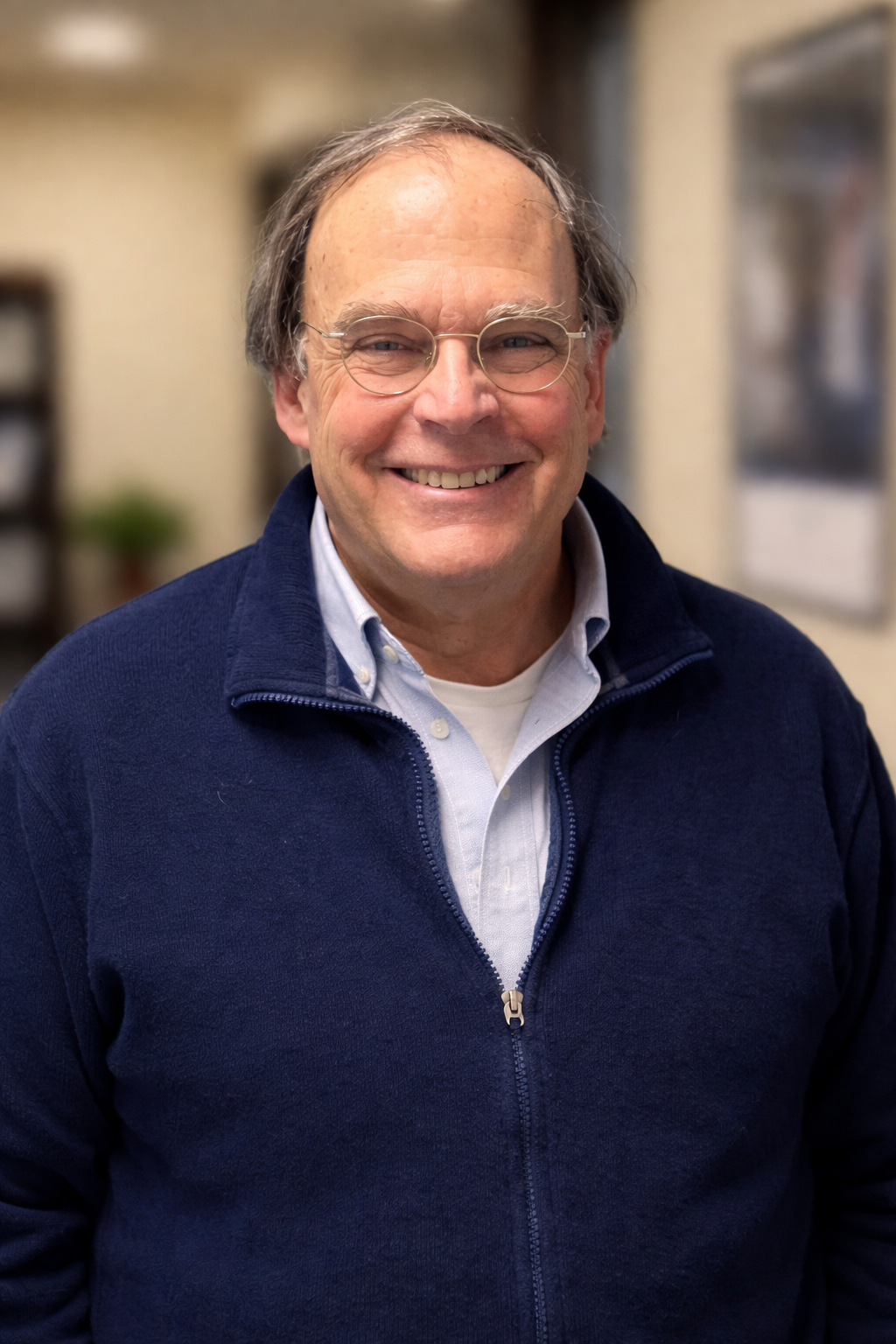 Smiling middle-aged man wearing glasses, a white shirt, and a navy blue zip-up sweater in an indoor setting.
