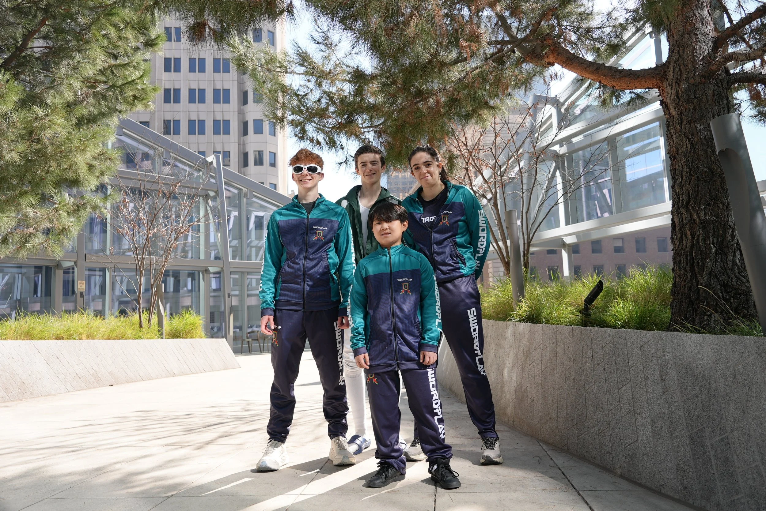 Four young people wearing matching sports outfits standing outdoors in front of a large building and trees, smiling for the camera.