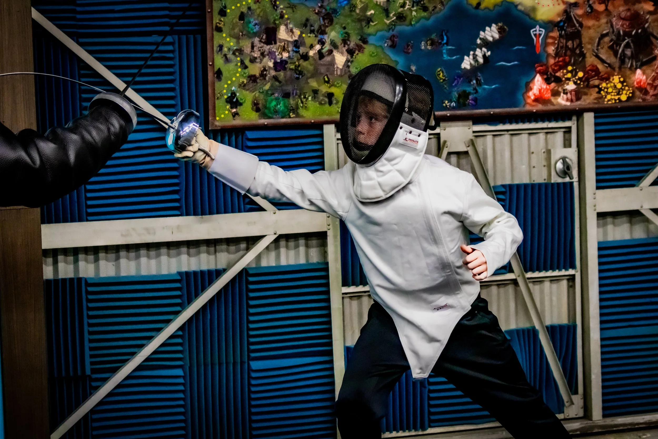 Two people wearing fencing gear practicing fencing in a training room, with wall-mounted training tools and a yellow training target in the background.
