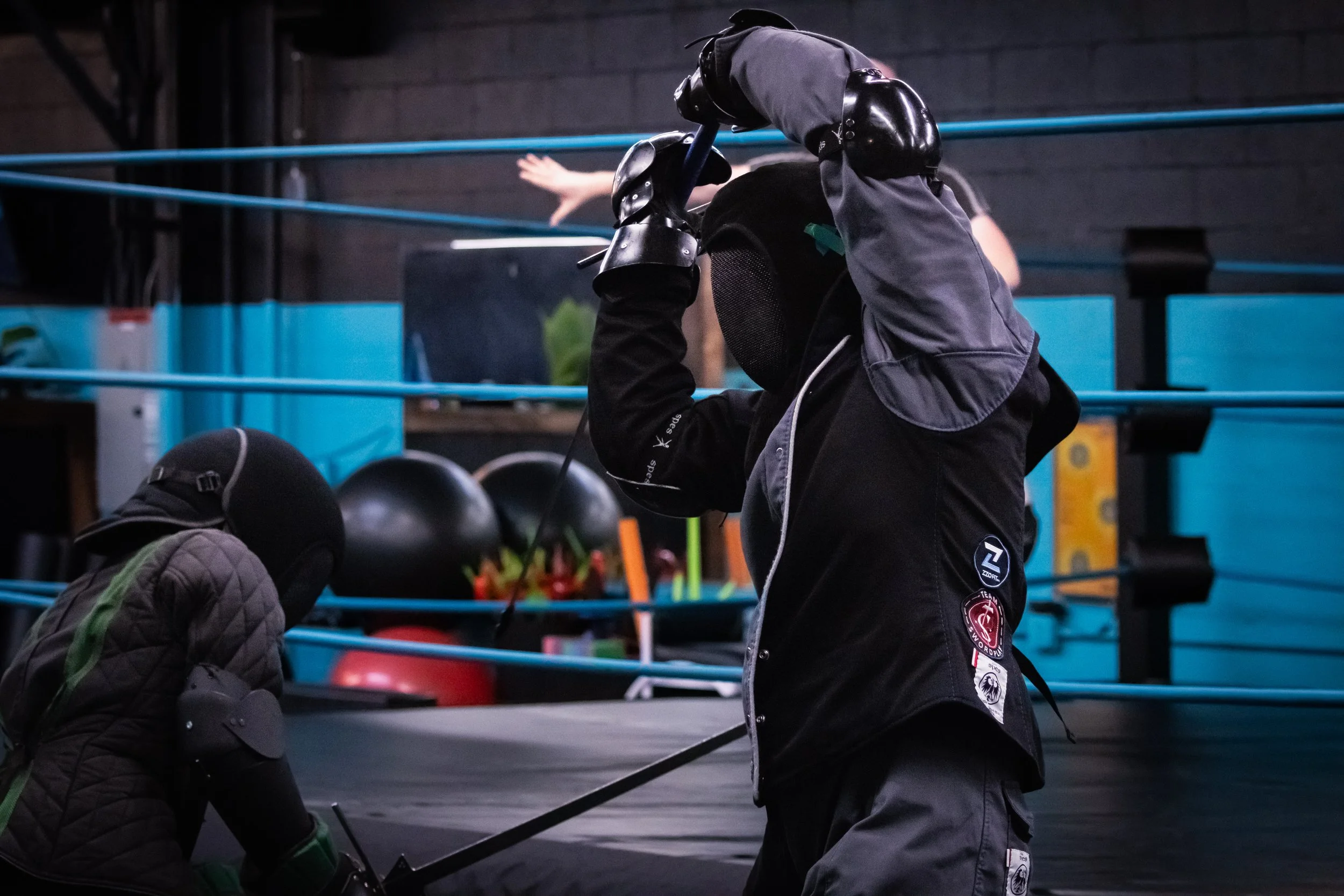 Two people practicing fencing with swords in a martial arts dojo, wearing protective gear including masks and padded clothing.