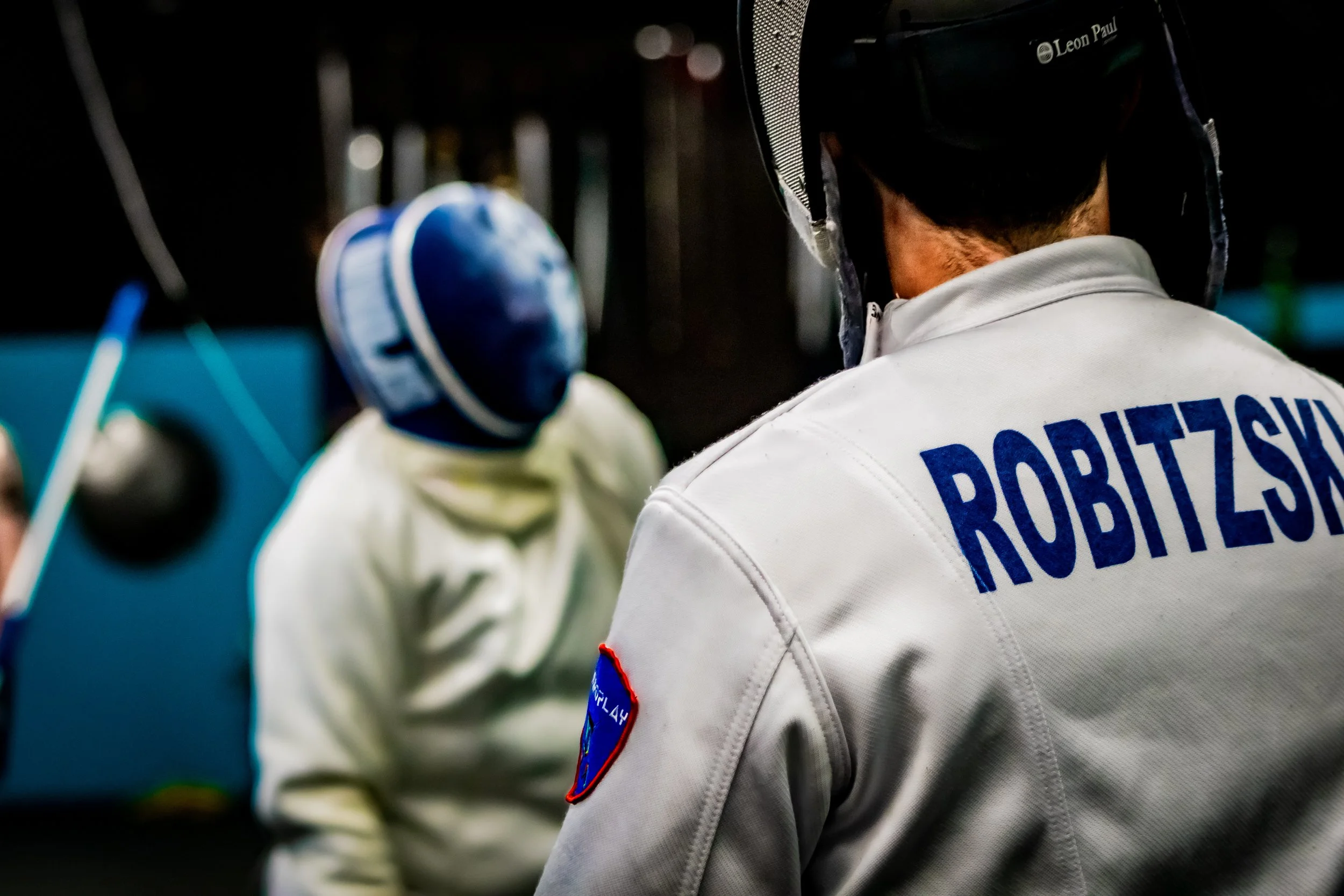 Group of fencers wearing protective gear and face masks, standing in a line in a fencing studio with swords mounted on the wall behind them.