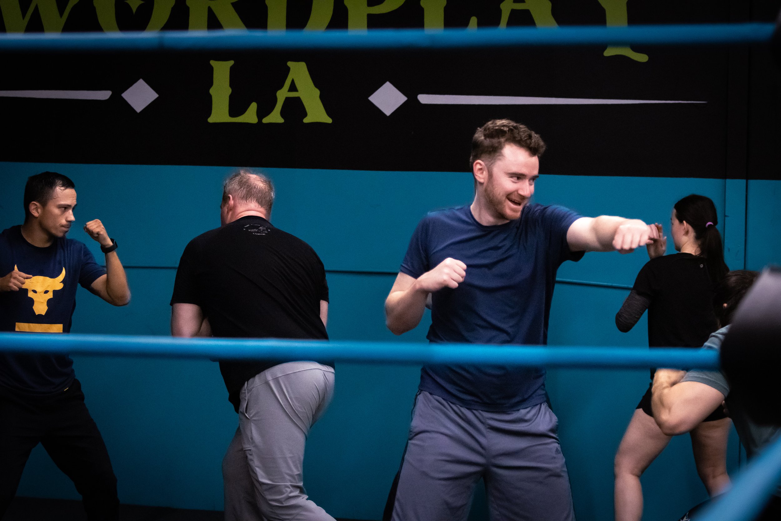 Three people practicing martial arts with padded sticks in a gym. One woman and one man appear to be sparring, while a third man is observing or instructing. The gym has martial arts weapons hanging on the wall and a large shield with decorative symbols.