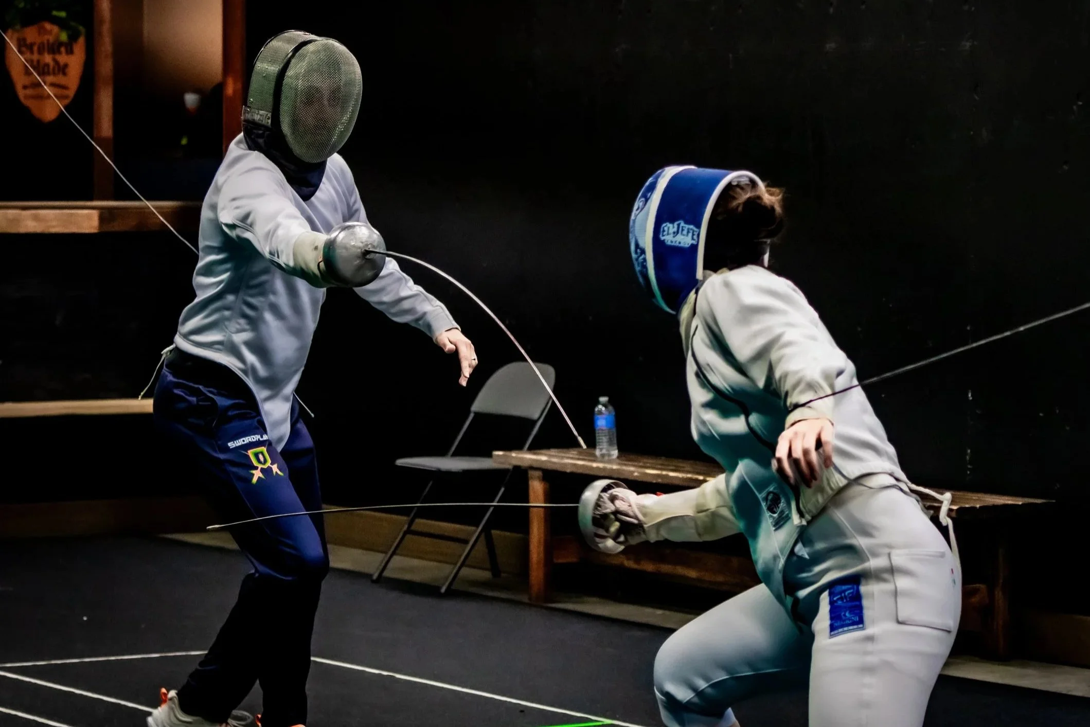 Two women fencing in protective gear sparring in a training facility with a group of people seated and watching in the background.