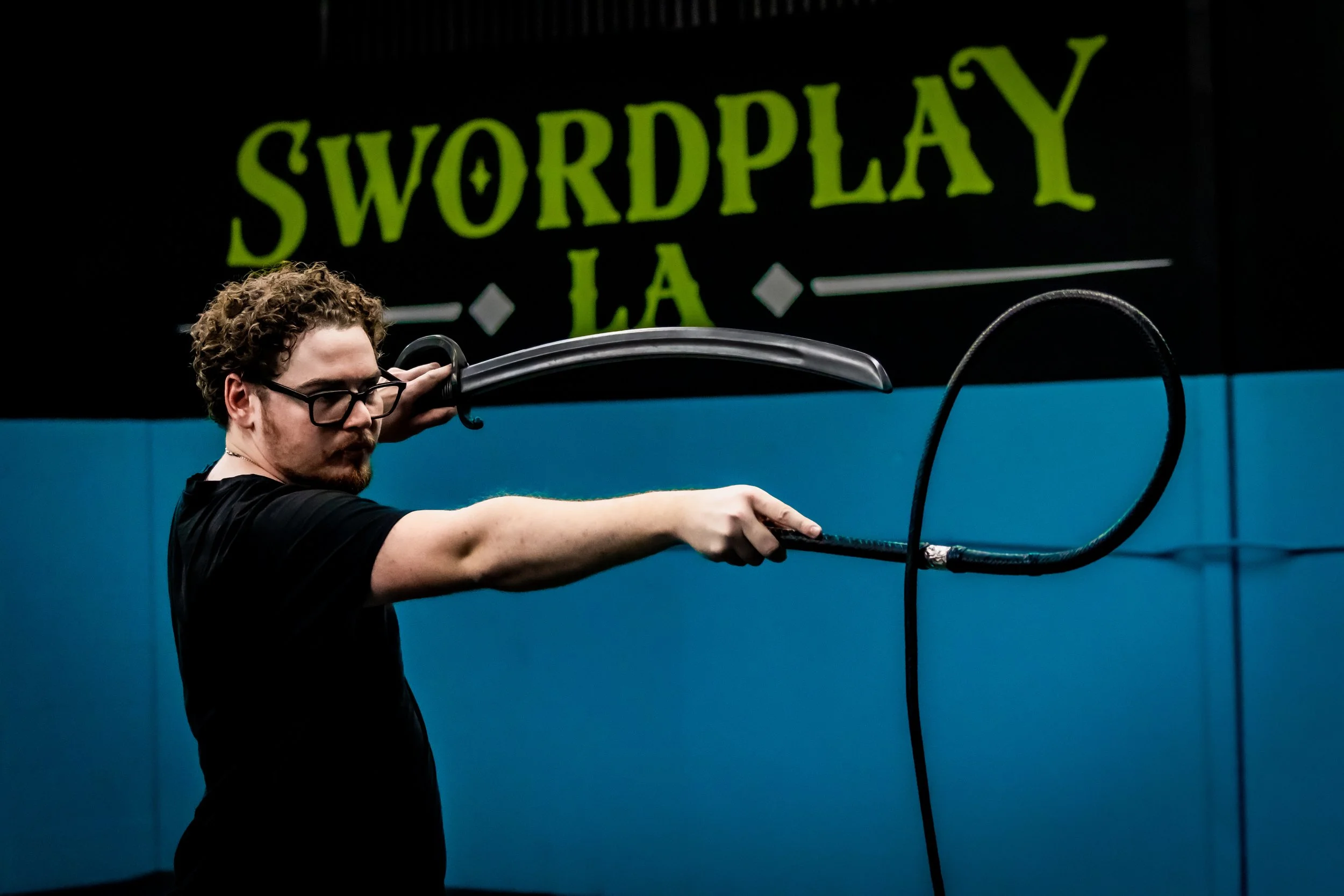 Three people practicing martial arts with padded sticks in a gym. One woman and one man appear to be sparring, while a third man is observing or instructing. The gym has martial arts weapons hanging on the wall and a large shield with decorative symbols.