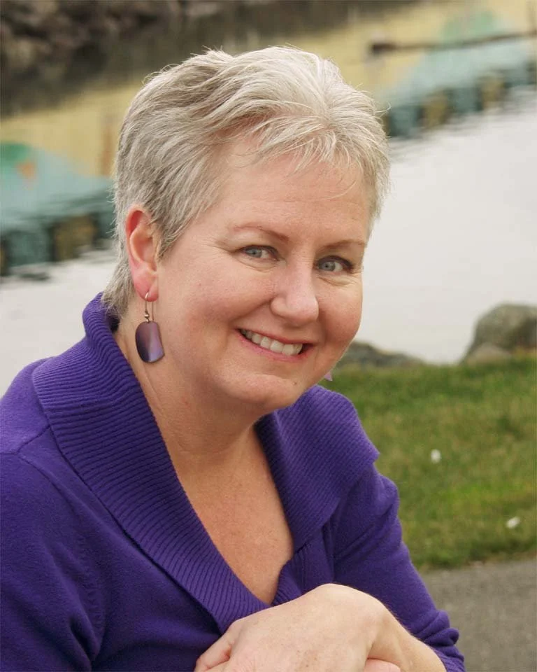 A smiling middle-aged woman with short gray hair, wearing purple earrings and a purple sweater, sitting outdoors near water with rocks and greenery in the background.