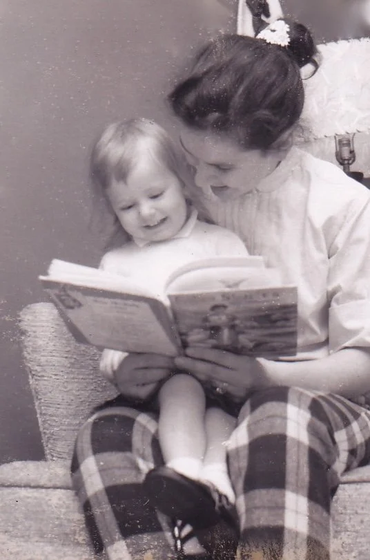 A woman and a young girl sitting on a chair reading a book together, smiling and enjoying a moment of shared reading.