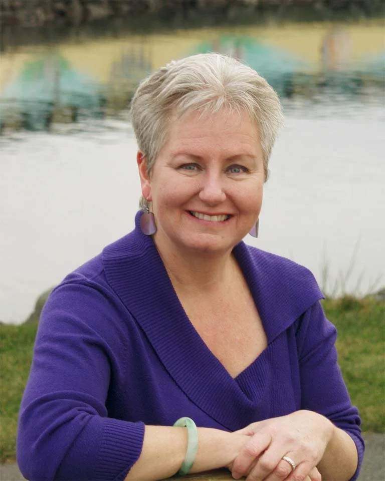 A smiling woman with short blonde hair, wearing a purple sweater and earrings, sitting outdoors near a body of water.