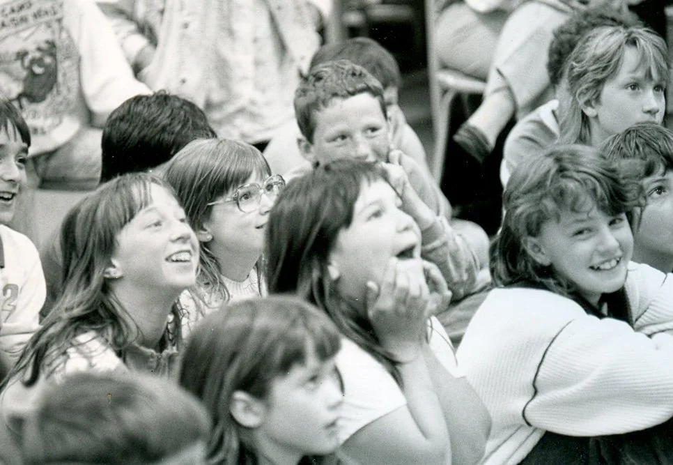 Children sitting in a classroom, smiling and laughing while watching a presentation or performance.