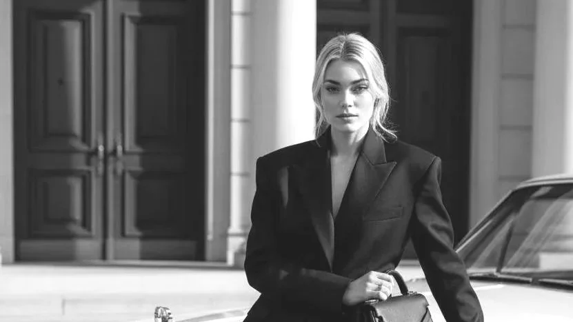 A woman with blonde hair and a serious expression wearing a black suit, standing beside a car in front of a building with large doors.