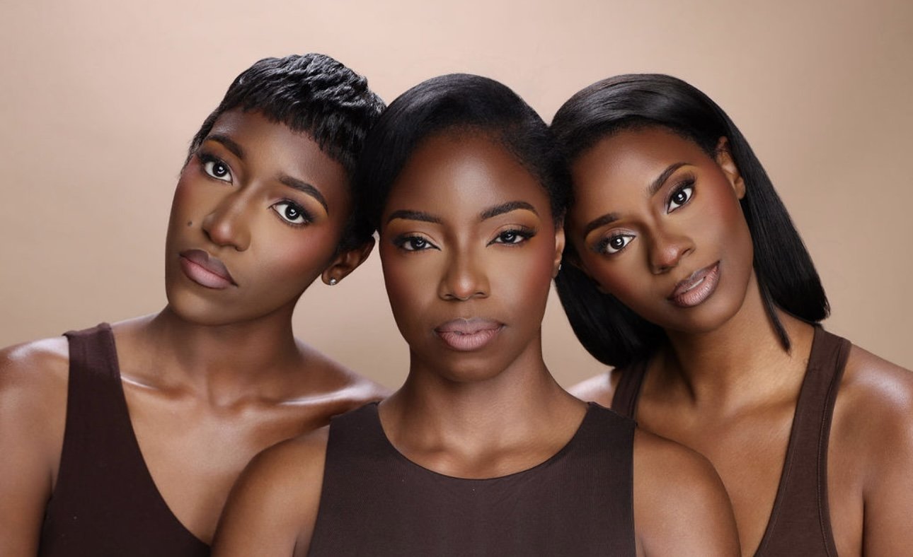 Three women of African descent with different hairstyles and makeup, all wearing brown tank tops, posing closely together against a neutral background.