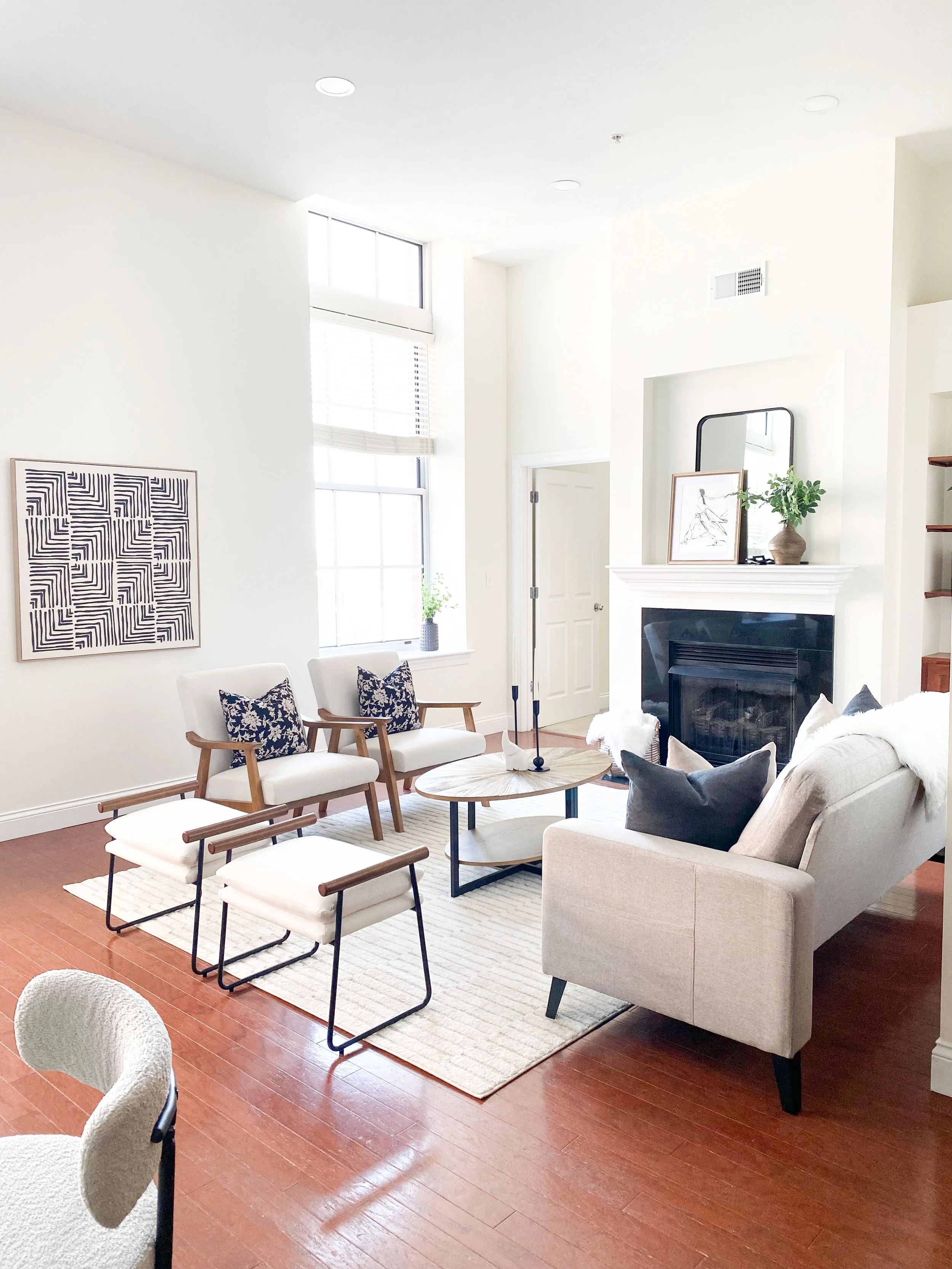 Modern living room with white walls, wooden flooring, a beige sofa with black and beige cushions, two matching white chairs with dark wood arms, a wooden coffee table, and a fireplace with a mirror and framed artwork on the mantelpiece. Large windows