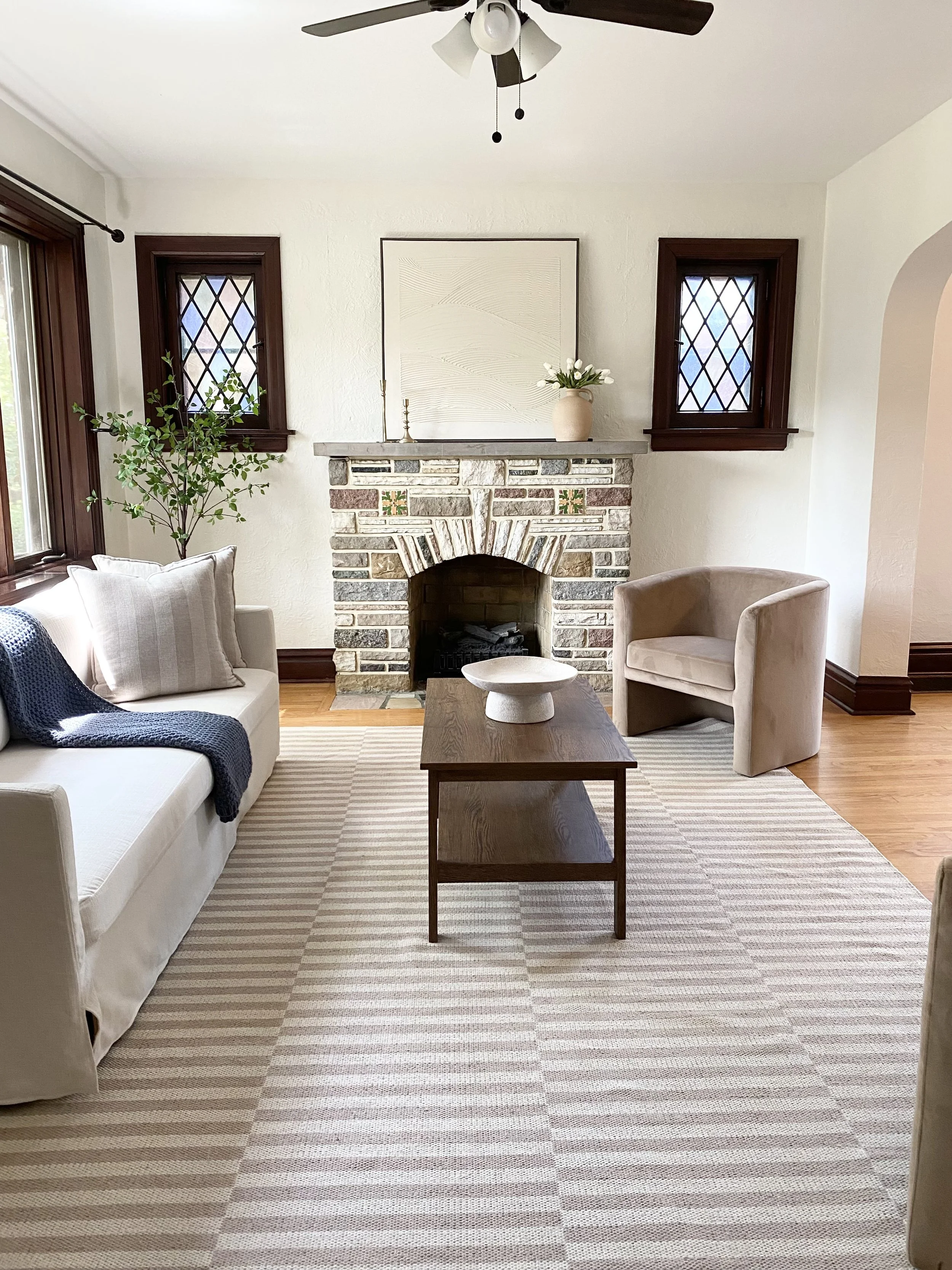 A cozy living room with a white sofa, beige armchair, wooden coffee table, stone fireplace, two small windows with diamond lattice panes, a large plant by the window, and minimal wall decor, including a white canvas and a vase with flowers on the man