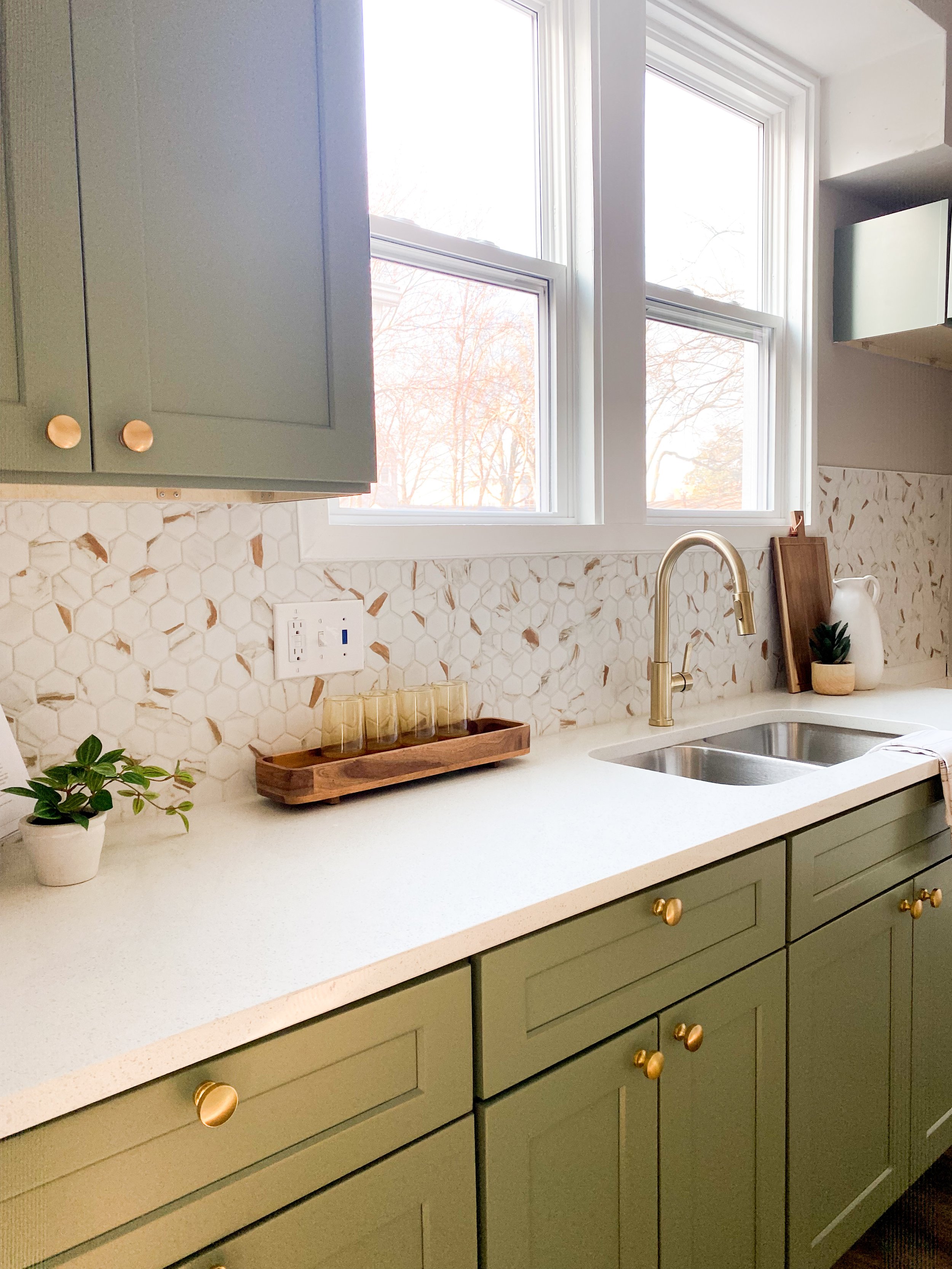 A kitchen countertop with green cabinets and gold knobs, a white marble backsplash, a double window, a gold faucet, a small potted plant, a wooden tray with glasses, and decorative items like a pitcher, cutting board, and a plant in a small pot.