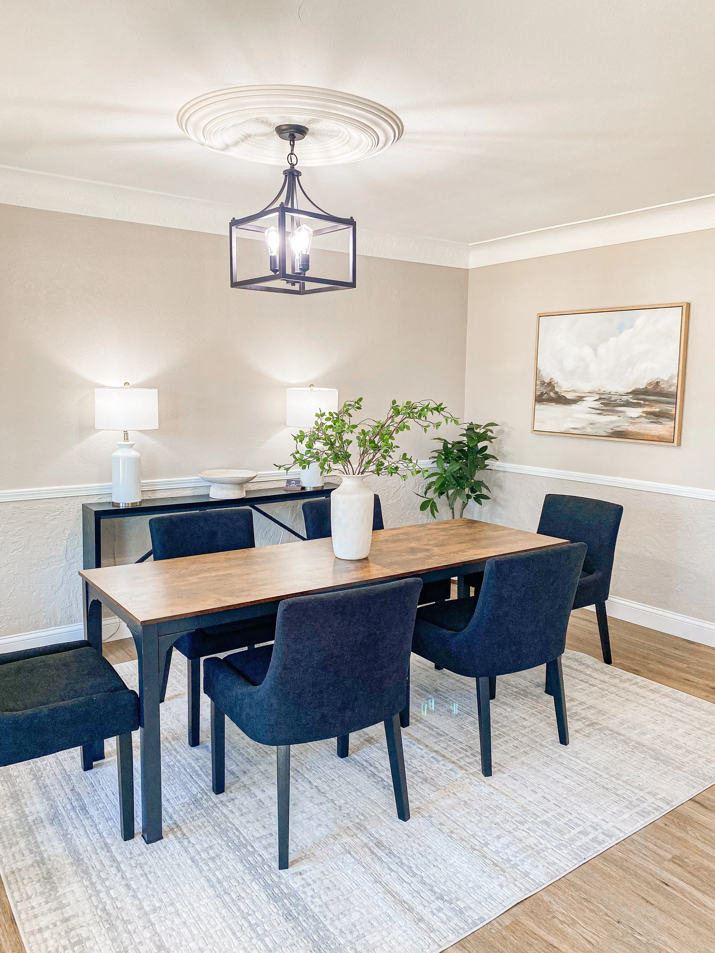 A dining room with a wooden table, six black chairs, a white rug, a black sideboard with two white lamps, two decorative objects, and a plant. A painting hangs on the beige wall. A black chandelier is on the ceiling.
