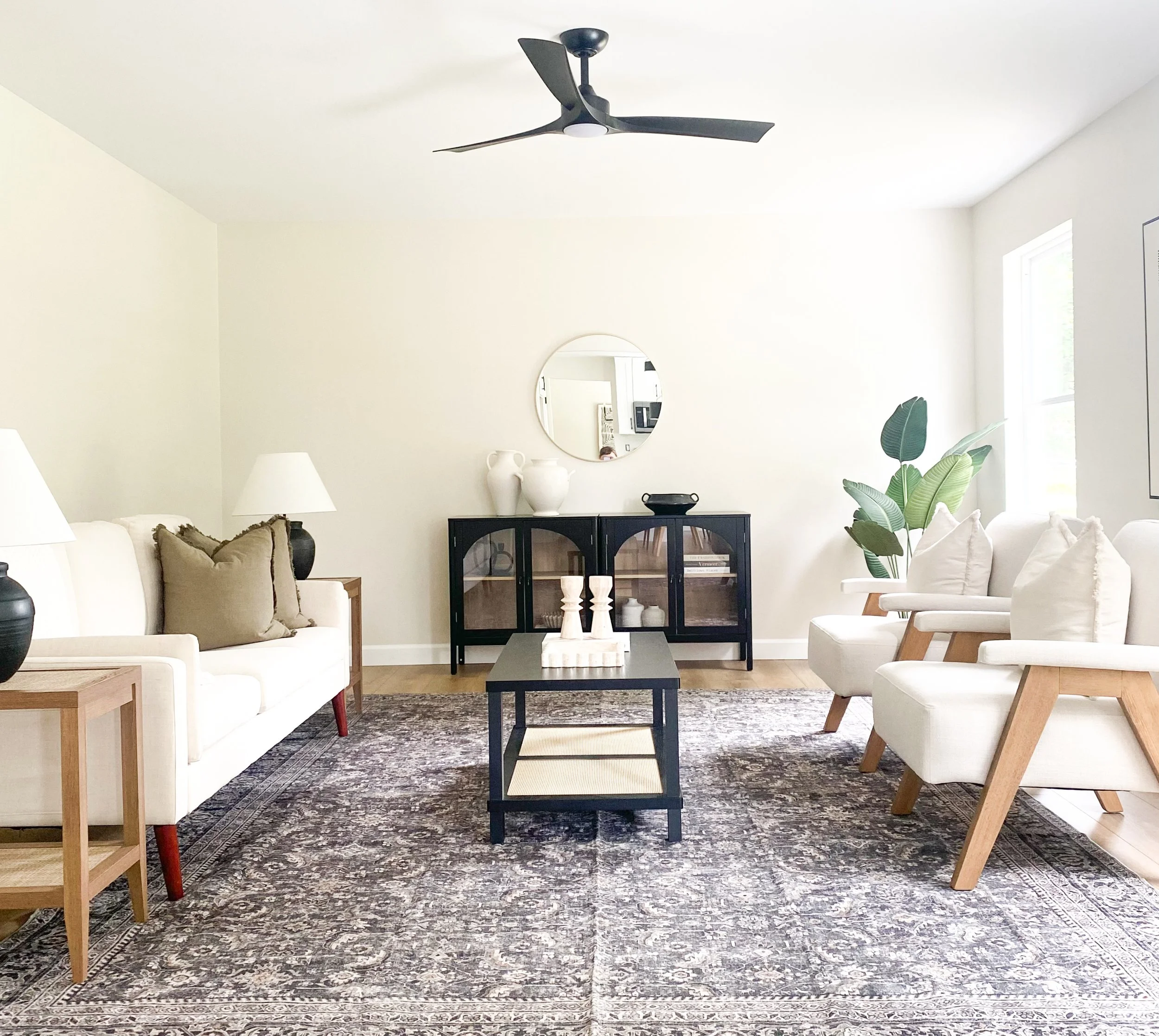 Living room with white walls, a black ceiling fan, a white sofa with brown pillows, two white armchairs, a black coffee table, a black cabinet with decorative vases, a mirror, a wooden side table with lamps, a large patterned rug, and a green housepl