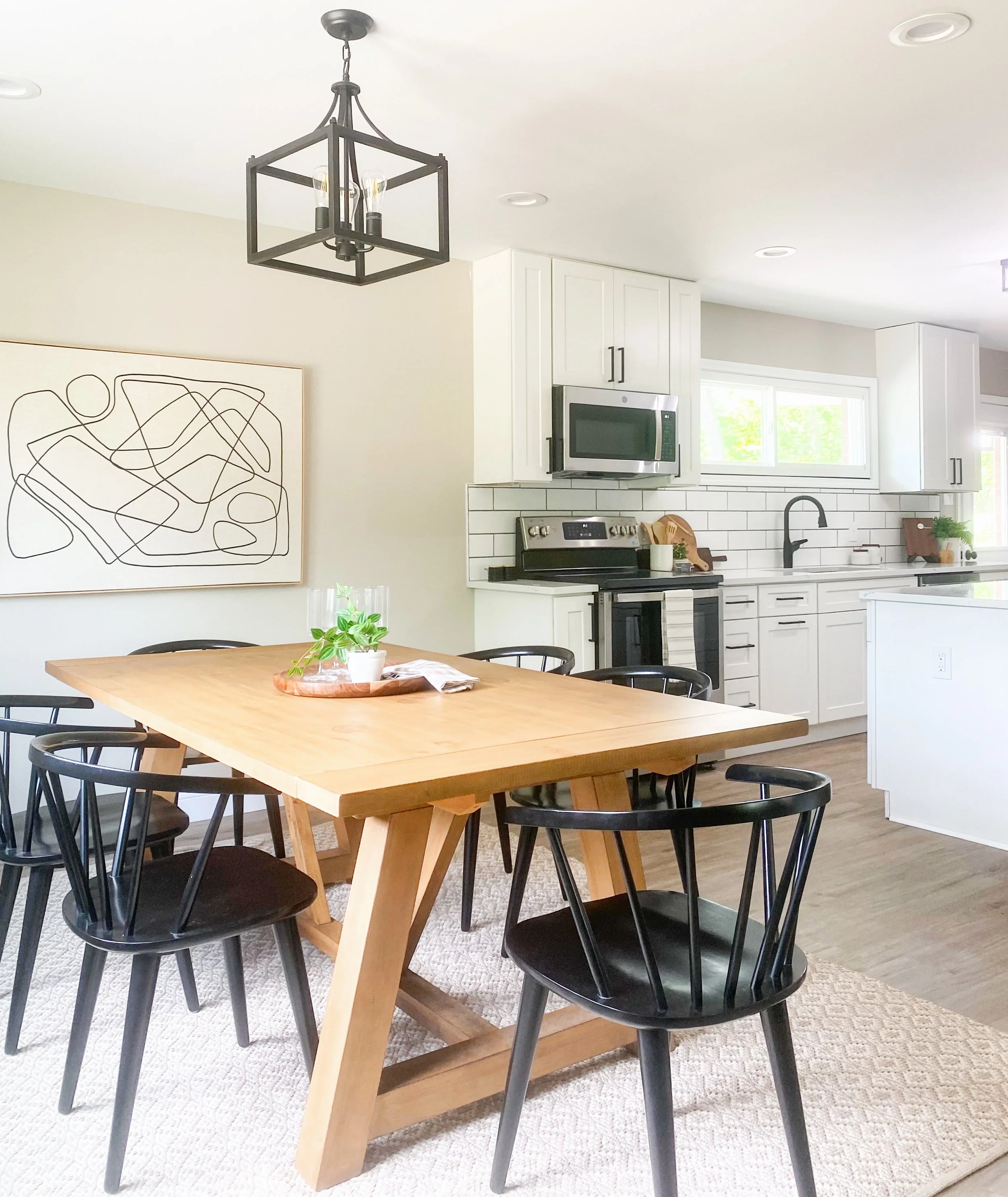 Modern kitchen and dining area with a wooden table, black chairs, white cabinetry, stainless steel stove and microwave, black faucet, window, and abstract artwork on the wall.