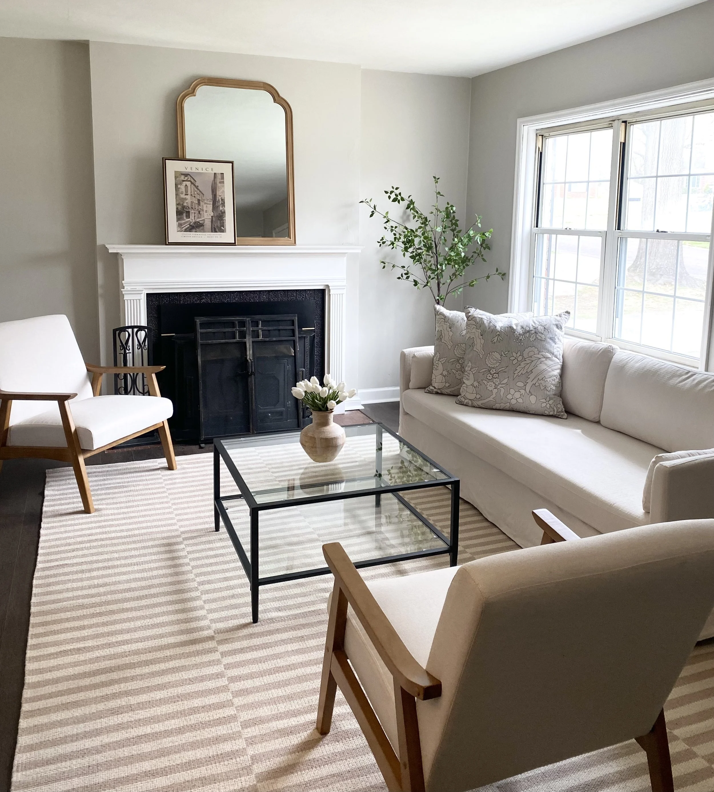 Living room with white sofa, armchairs with wooden arms, a glass coffee table with a vase of white flowers, a fireplace with a mirror and framed picture, and a large window bringing in natural light.