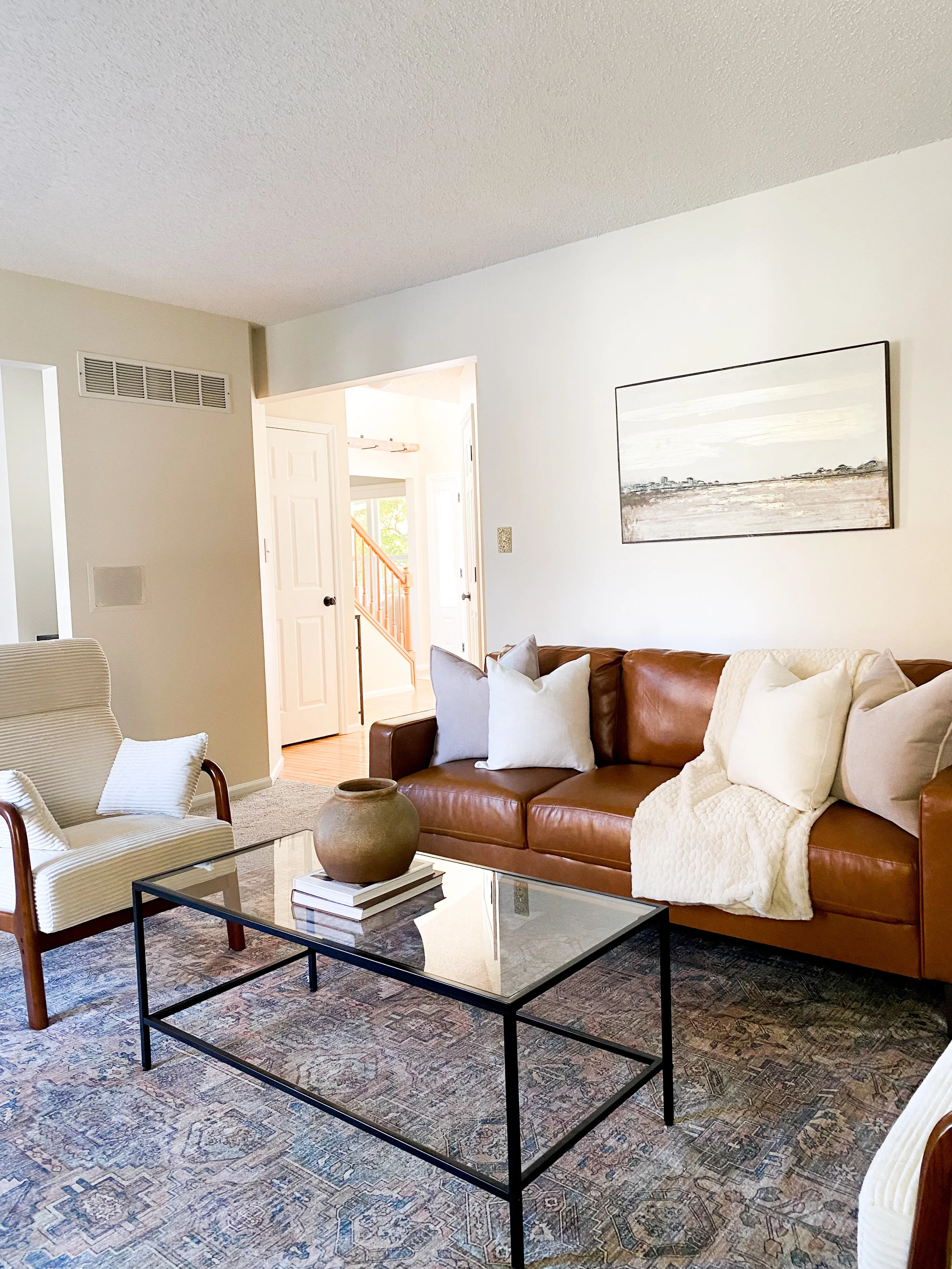 Living room with brown leather sofa, cream armchair, glass coffee table, decorative vase on books, abstract landscape art on white wall, patterned rug, and open doorway to entryway.