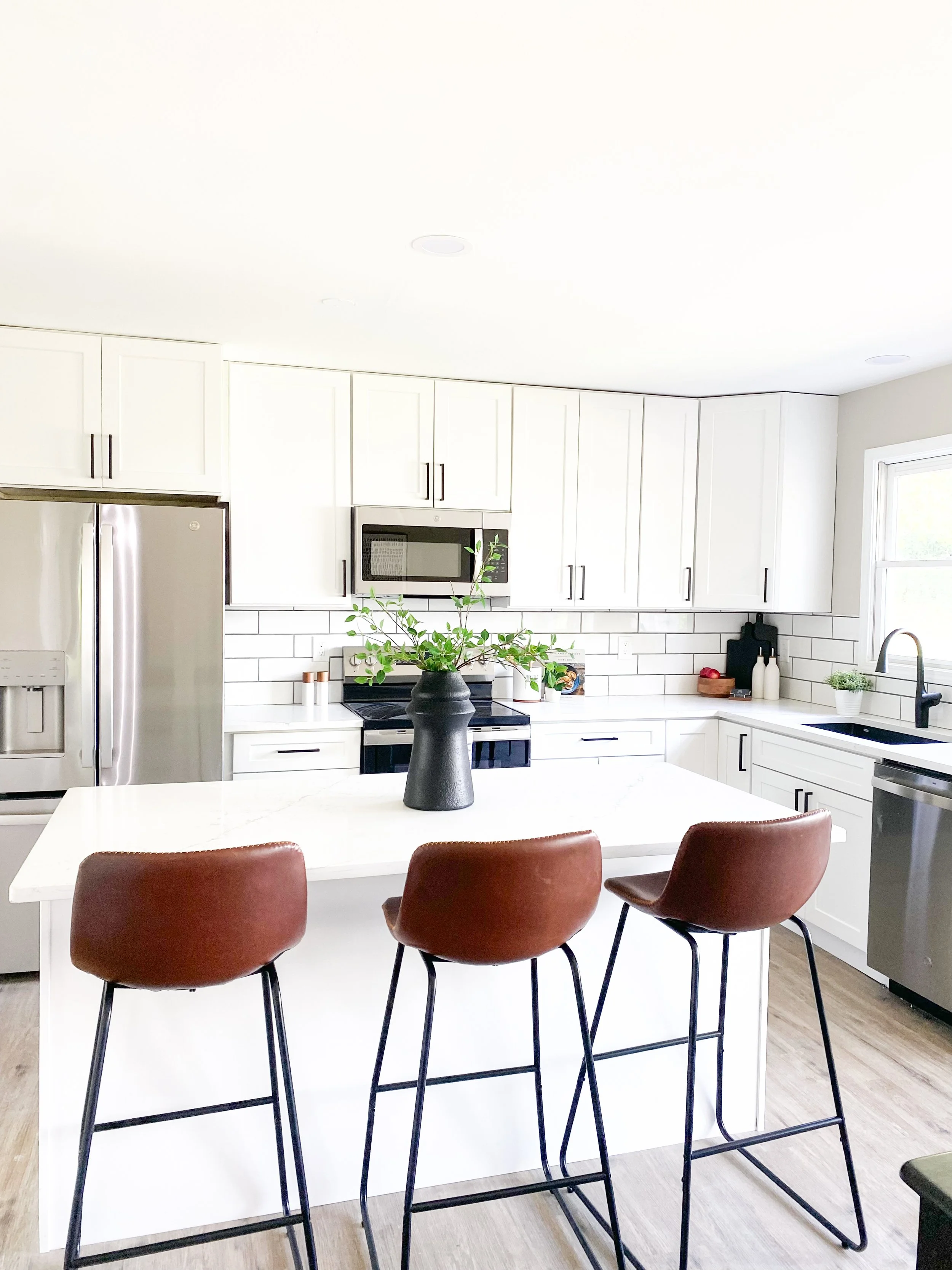 Modern kitchen with white cabinets, a marble island with three brown chairs, stainless steel appliances, a black vase with green plant on the island, white subway tile backsplash, and a window above the sink.