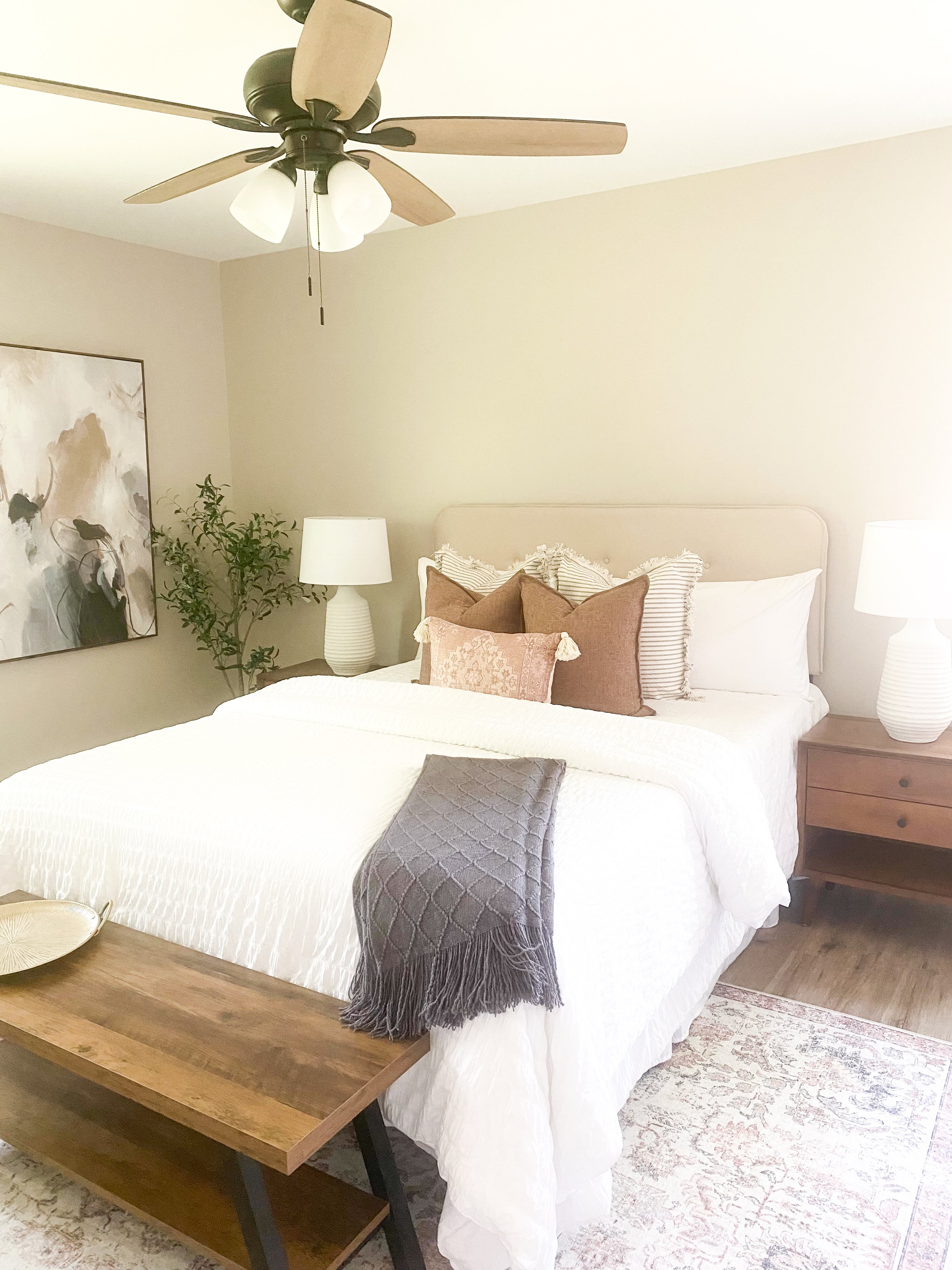 A cozy bedroom with a large bed covered in white bedding, decorated with multiple pillows, including striped and solid brown and pink ones. A gray throw blanket with tassels is draped over the foot of the bed. There are nightstands with white lamps o