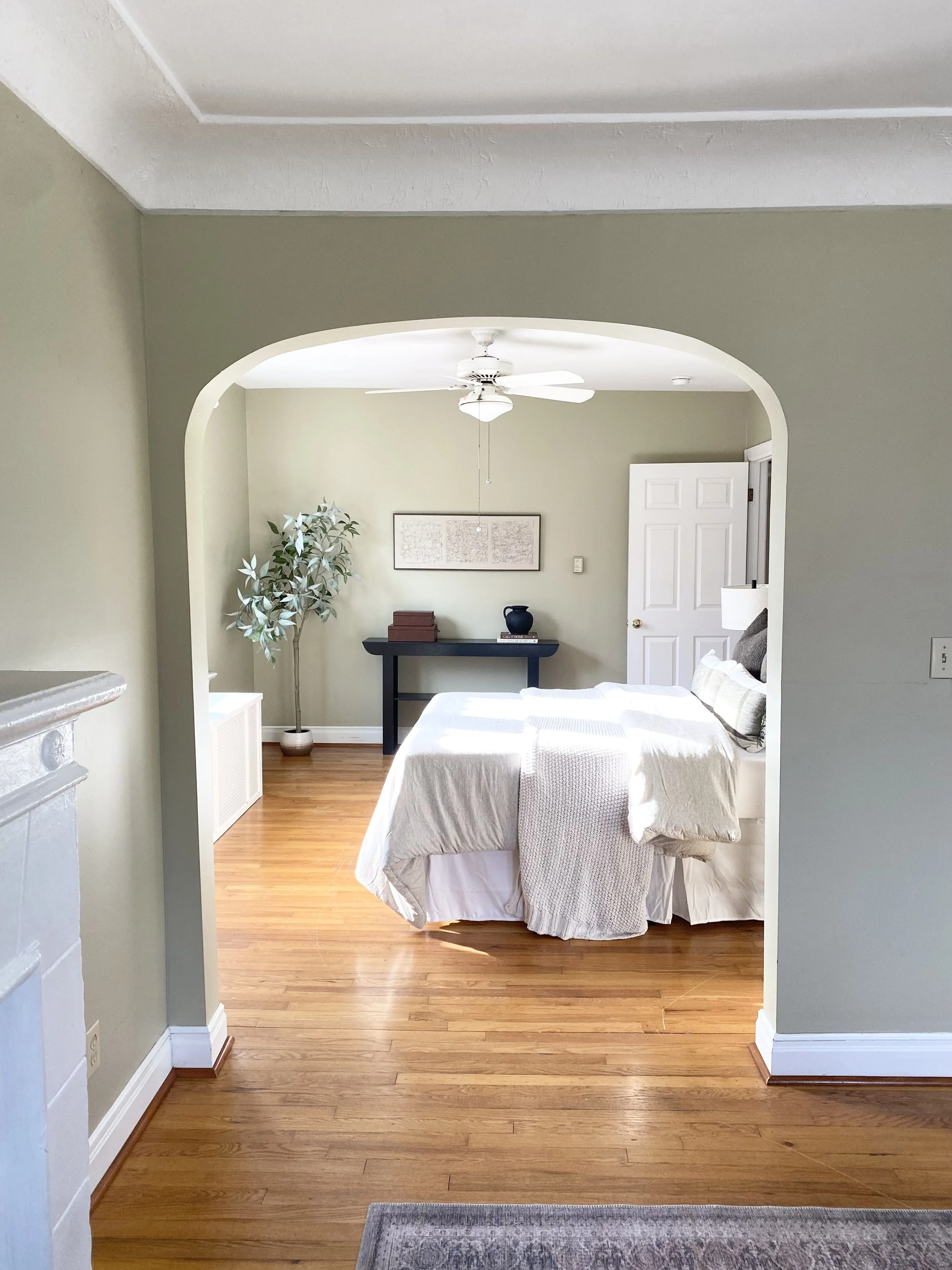 A bedroom with a bed covered in white bedding, hardwood floors, a ceiling fan, a white door, a black console table with decorative items, a framed map on the wall, a large potted plant, and a window allowing natural light.
