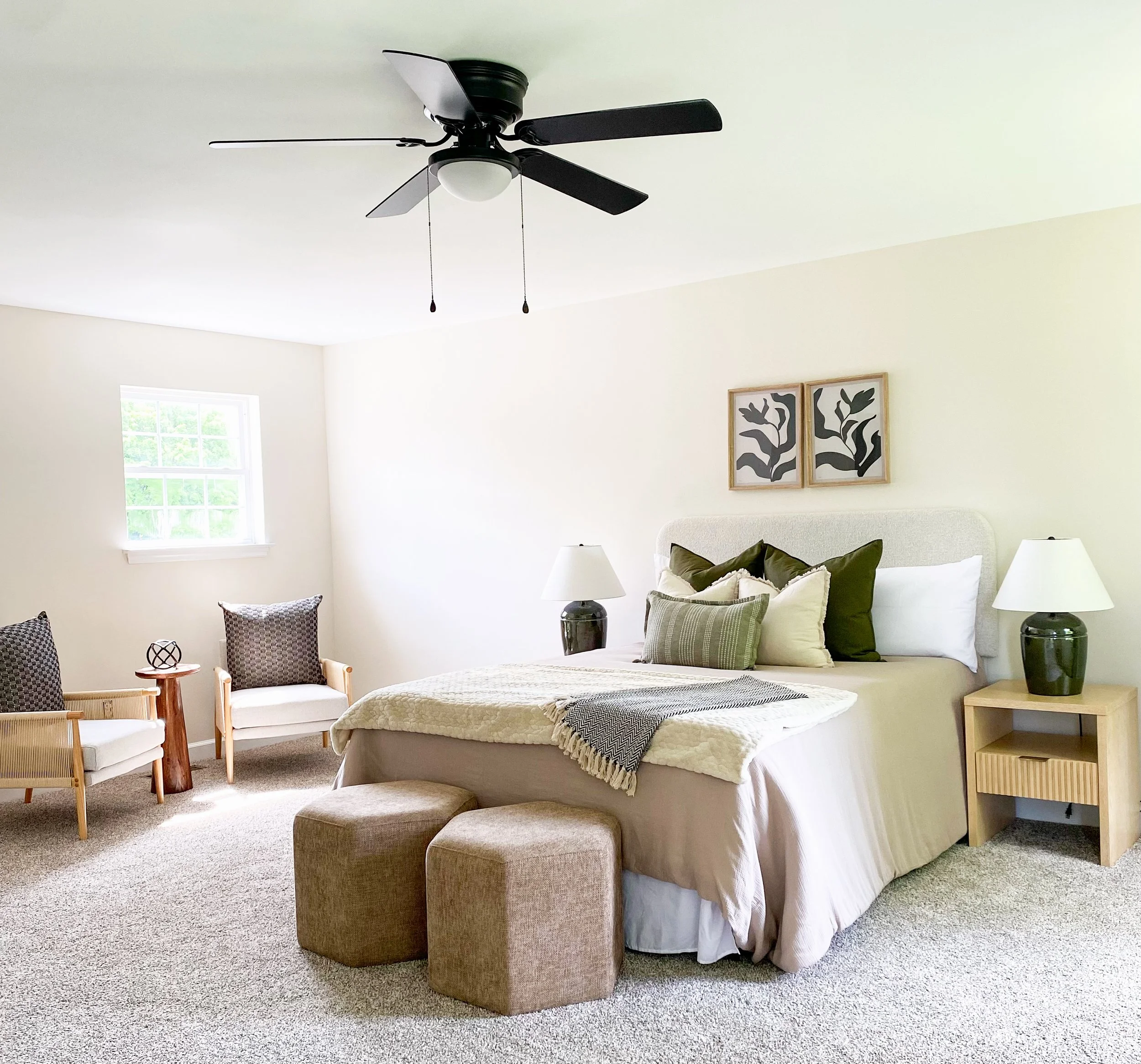 A bedroom with a large bed, two bedside tables with lamps, two square ottomans at the foot of the bed, two decorative pillows, a knitted blanket on the bed, and two framed pictures of abstract plant designs on the wall. In the corner, there are two c