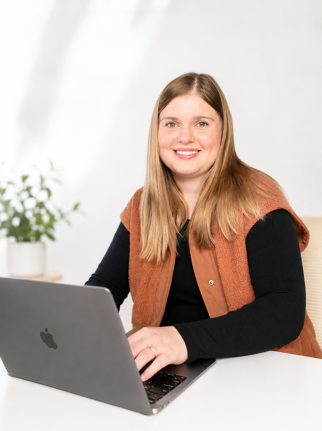 A woman with long blonde hair smiling while working on a laptop at a wooden table in a room with shelves and supplies in the background.