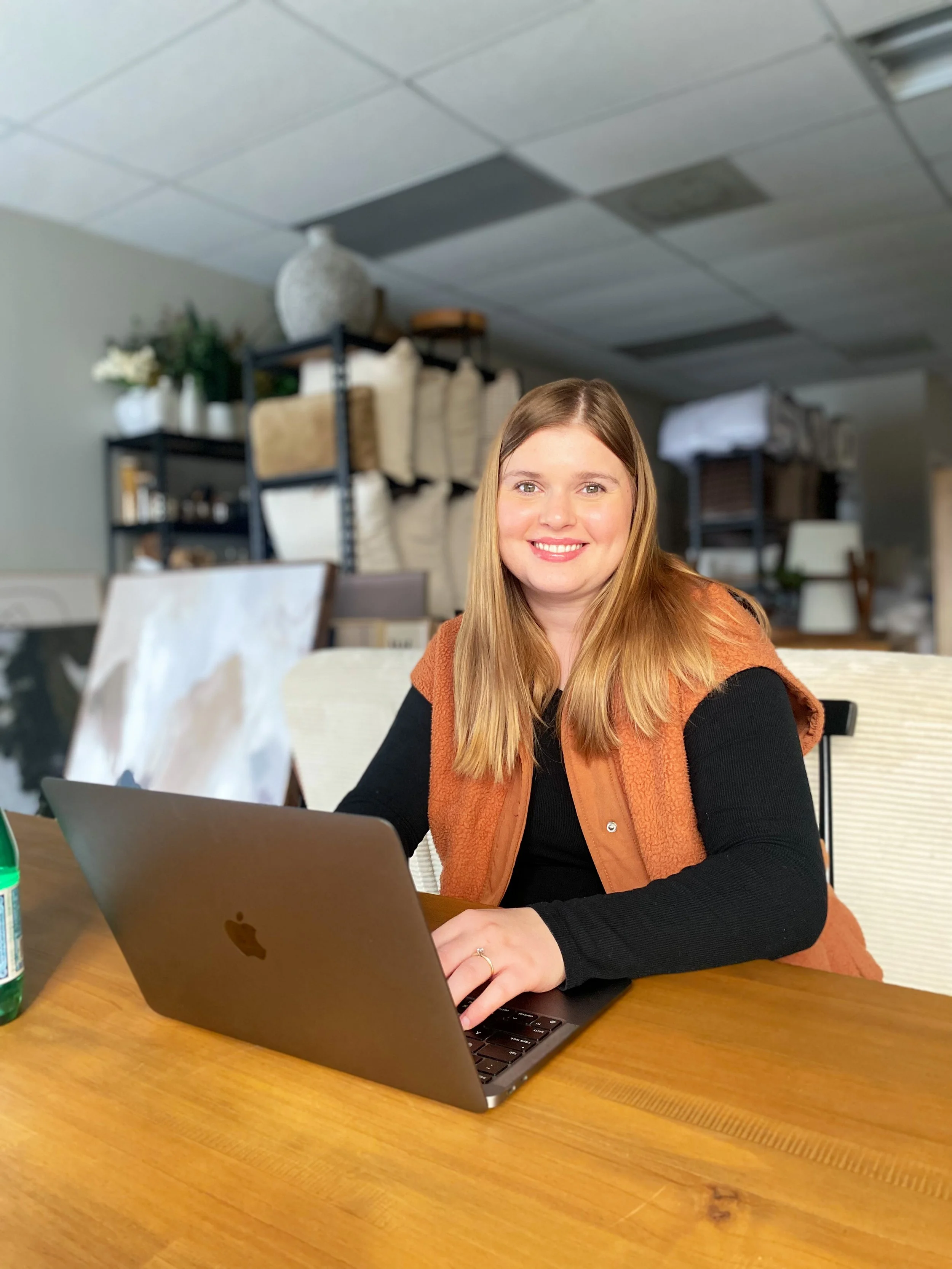 A woman with long blonde hair smiling while working on a laptop at a wooden table in a room with shelves and supplies in the background.