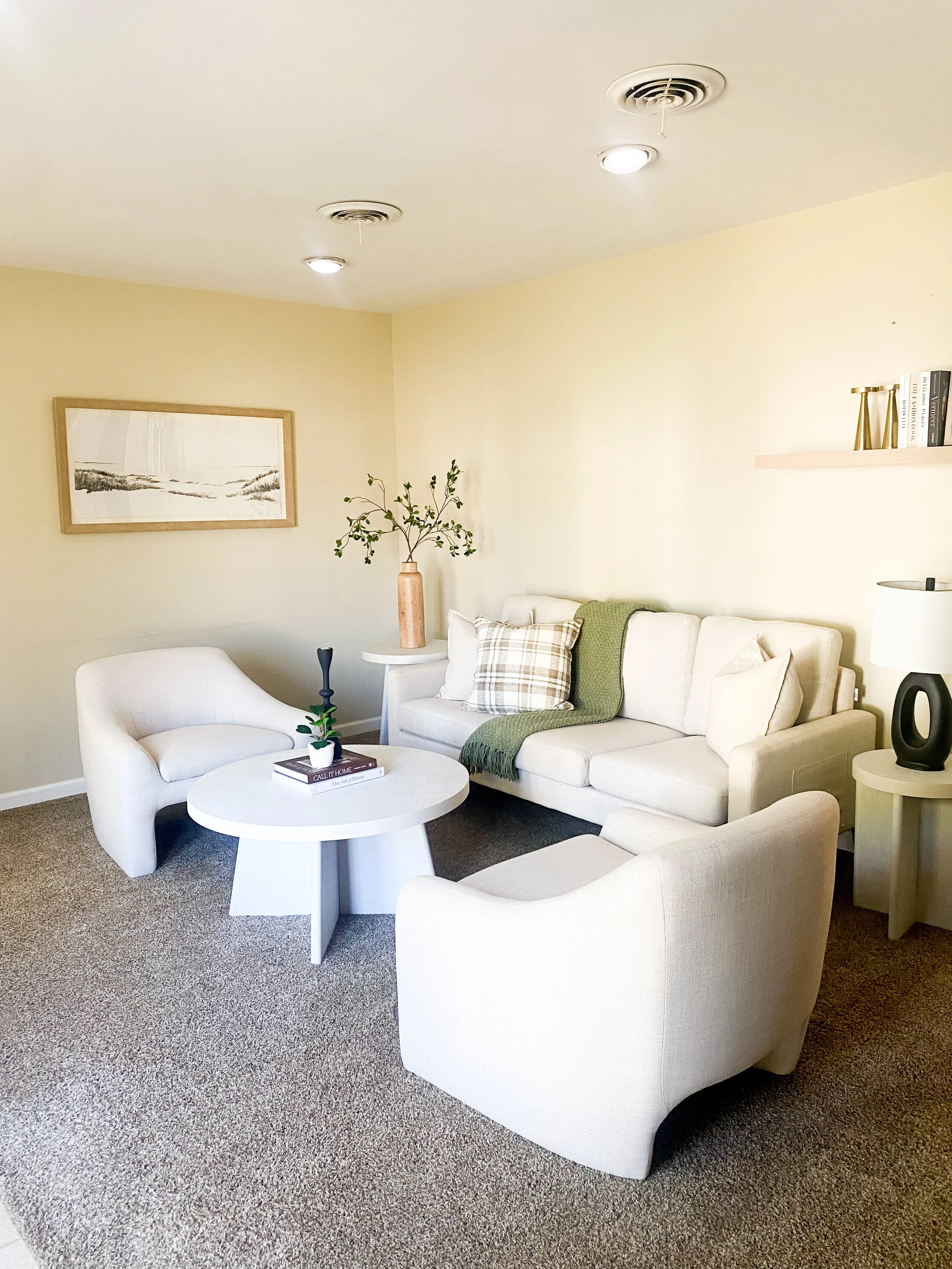 Living room with beige sofa and matching armchairs, a white round coffee table, decorative pillows, a side table with a table lamp, a framed artwork, a vase with branches, and a beige carpet.
