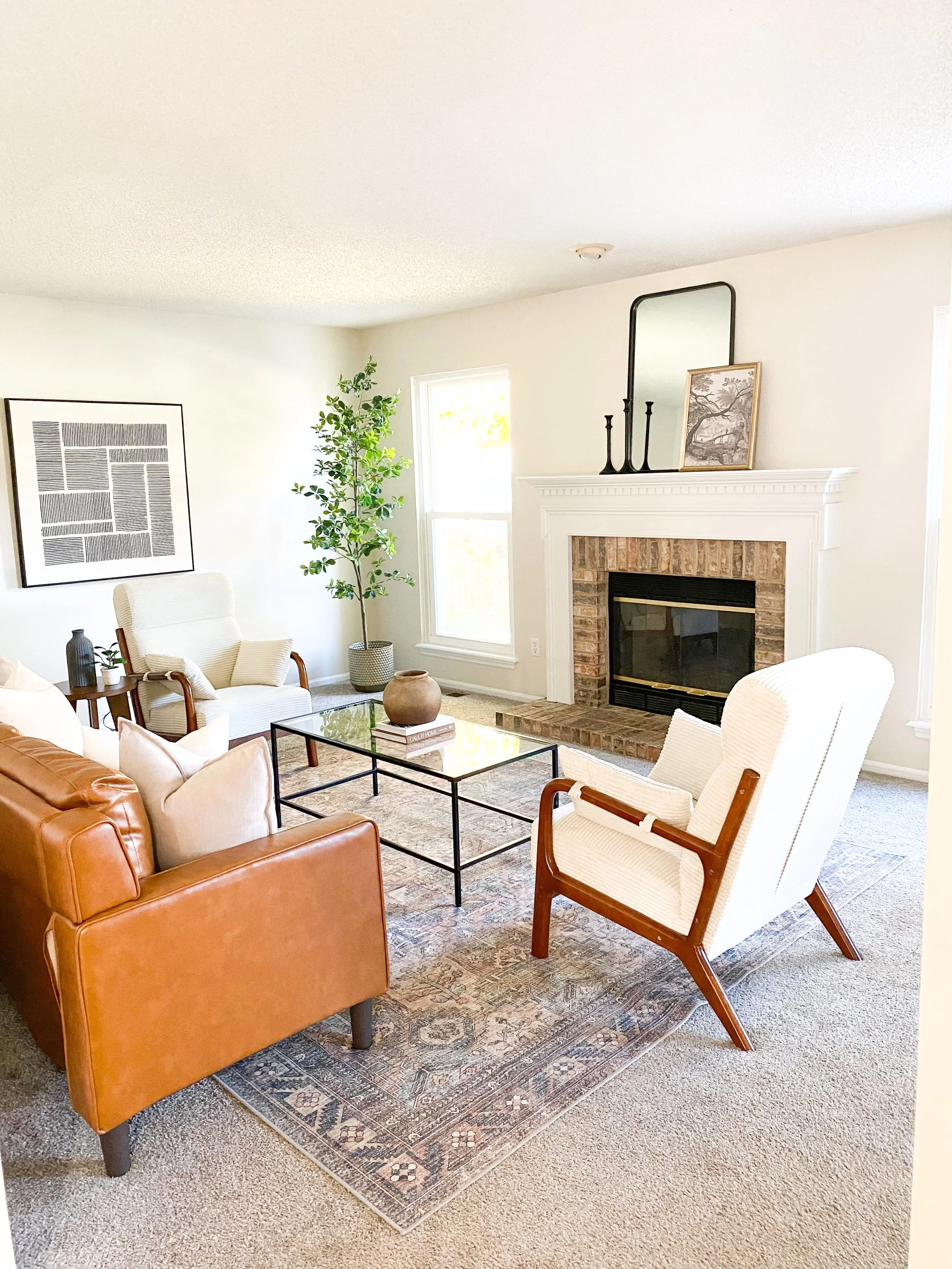 Bright living room with white walls, a brick fireplace, and natural light, featuring a tan leather sofa, white armchairs, a glass coffee table, a potted plant, and decorative items on the mantel.