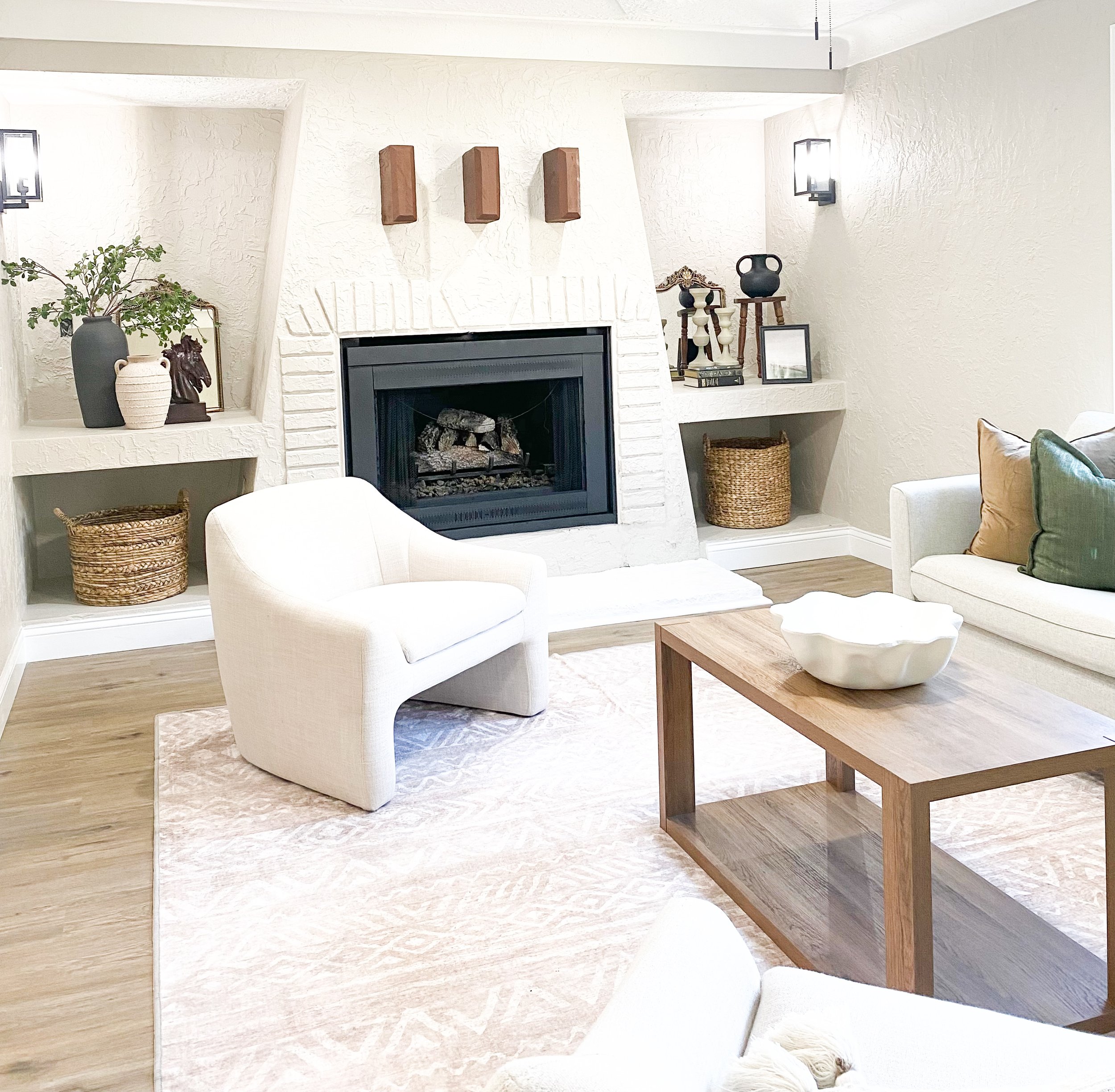 A cozy living room corner with a white upholstered armchair, a wooden coffee table with a white ceramic bowl, a white sofa with beige and green pillows, a white textured fireplace with decorations on the shelf and wall, and a light-colored area rug o