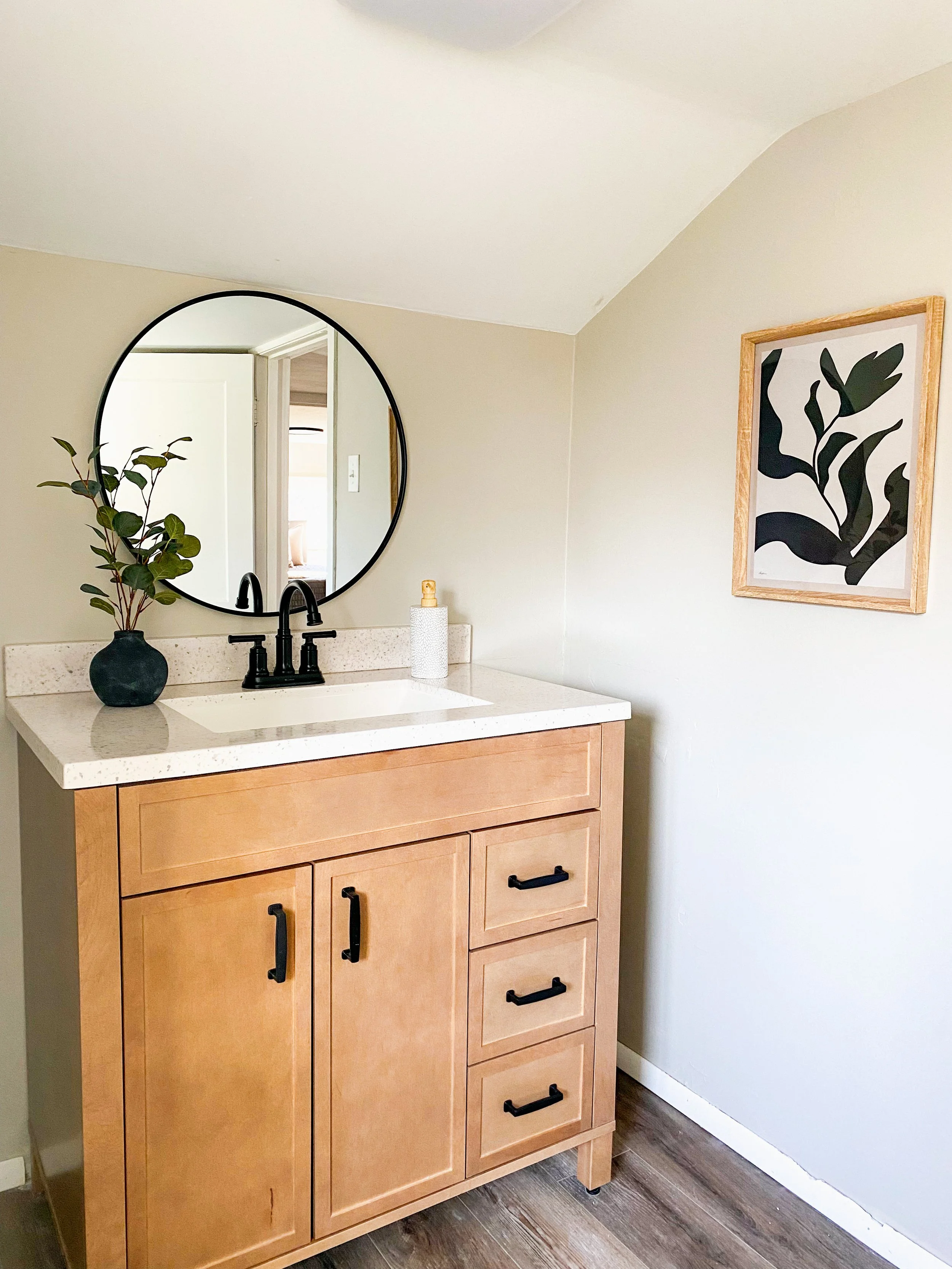 Bathroom vanity with a round mirror, a black faucet, a small black vase with greenery, and a framed black and white abstract art on the wall.