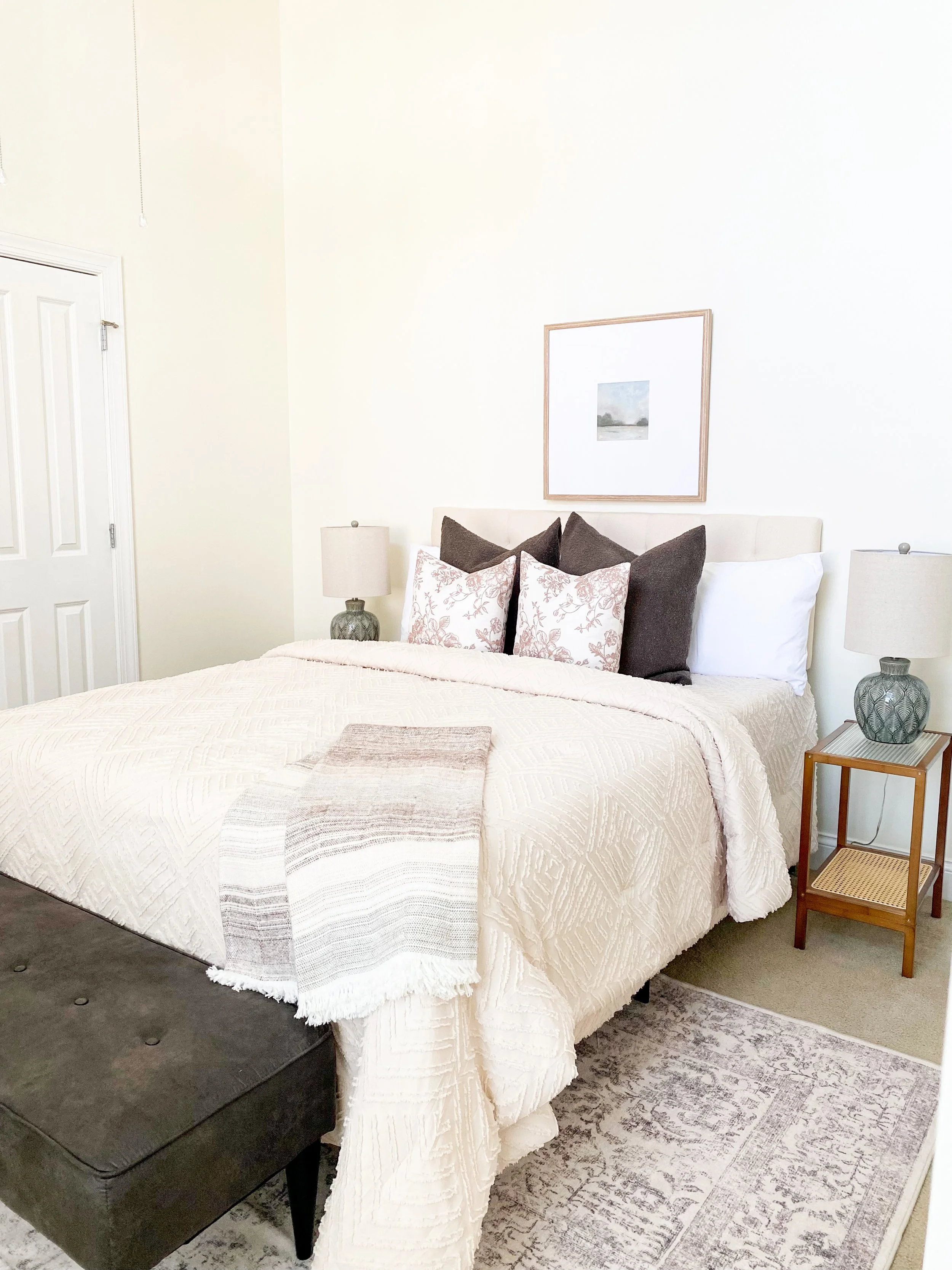 A cozy bedroom with a cream-colored bed, artwork above, nightstands with lamps, a black bench at the foot, and a patterned rug.