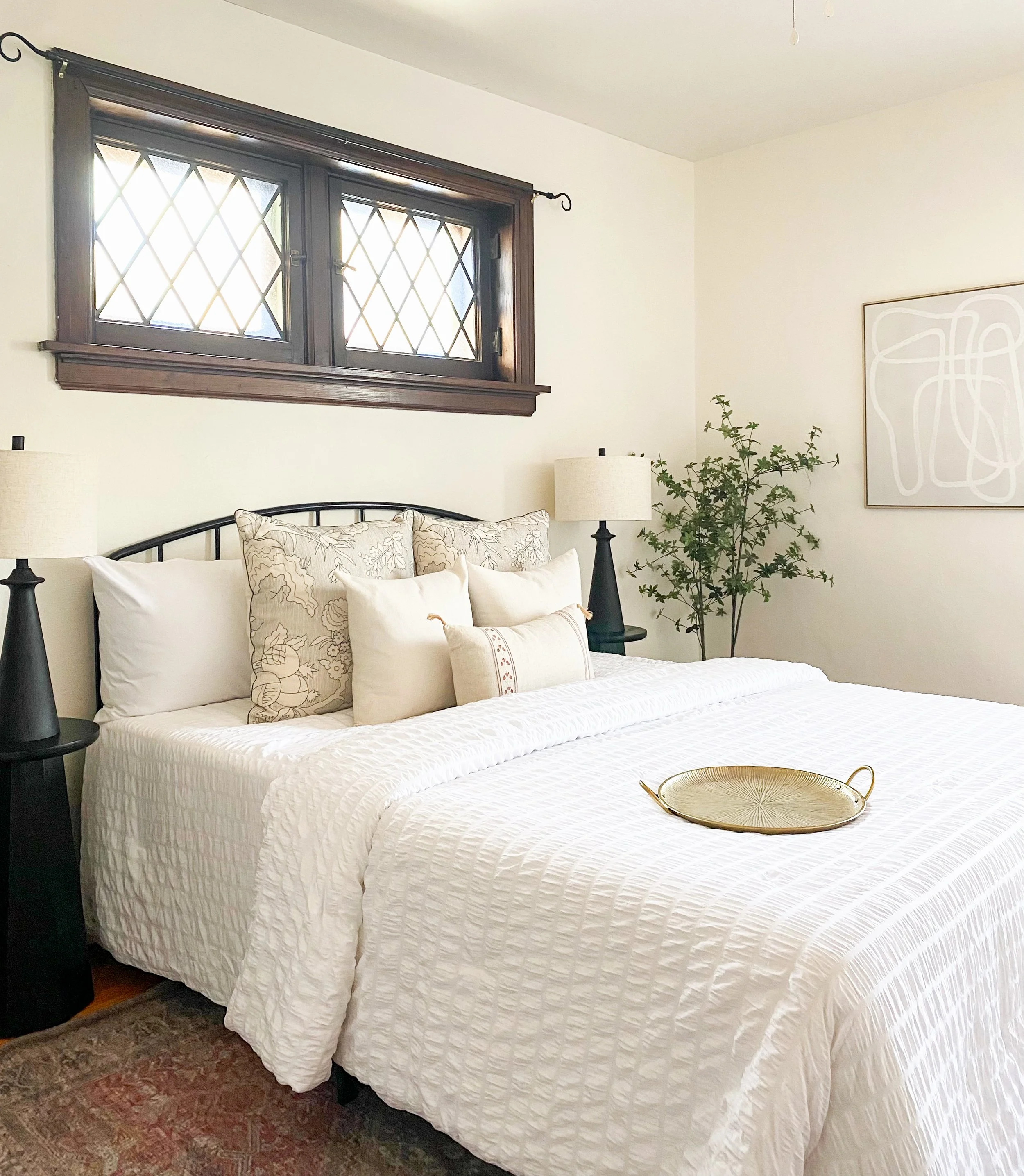 A neatly made bed with white bedding, multiple pillows, and a decorative tray on top. Two black nightstands with beige lamps flank the bed. A potted plant and artwork are on the wall behind, and a window with diamond-shaped panes is above the bed.