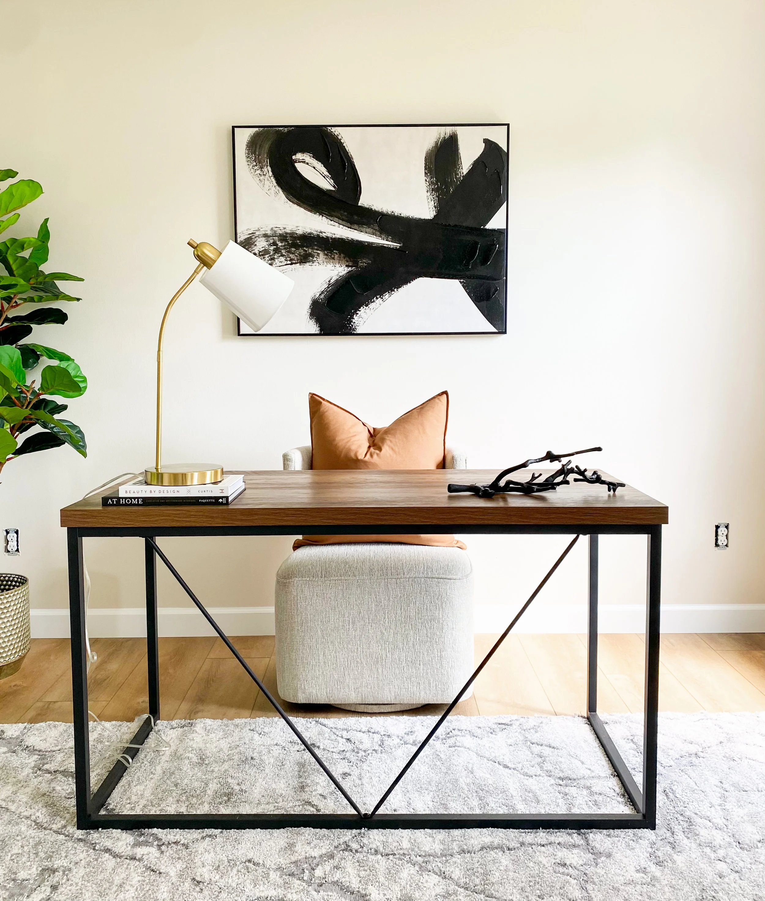A modern home office with a wooden desk, a white plush chair, a brown cushion, a bookshelf with books, a table lamp, a branch decor item, abstract black and white artwork on the wall, a potted plant, and a rug.