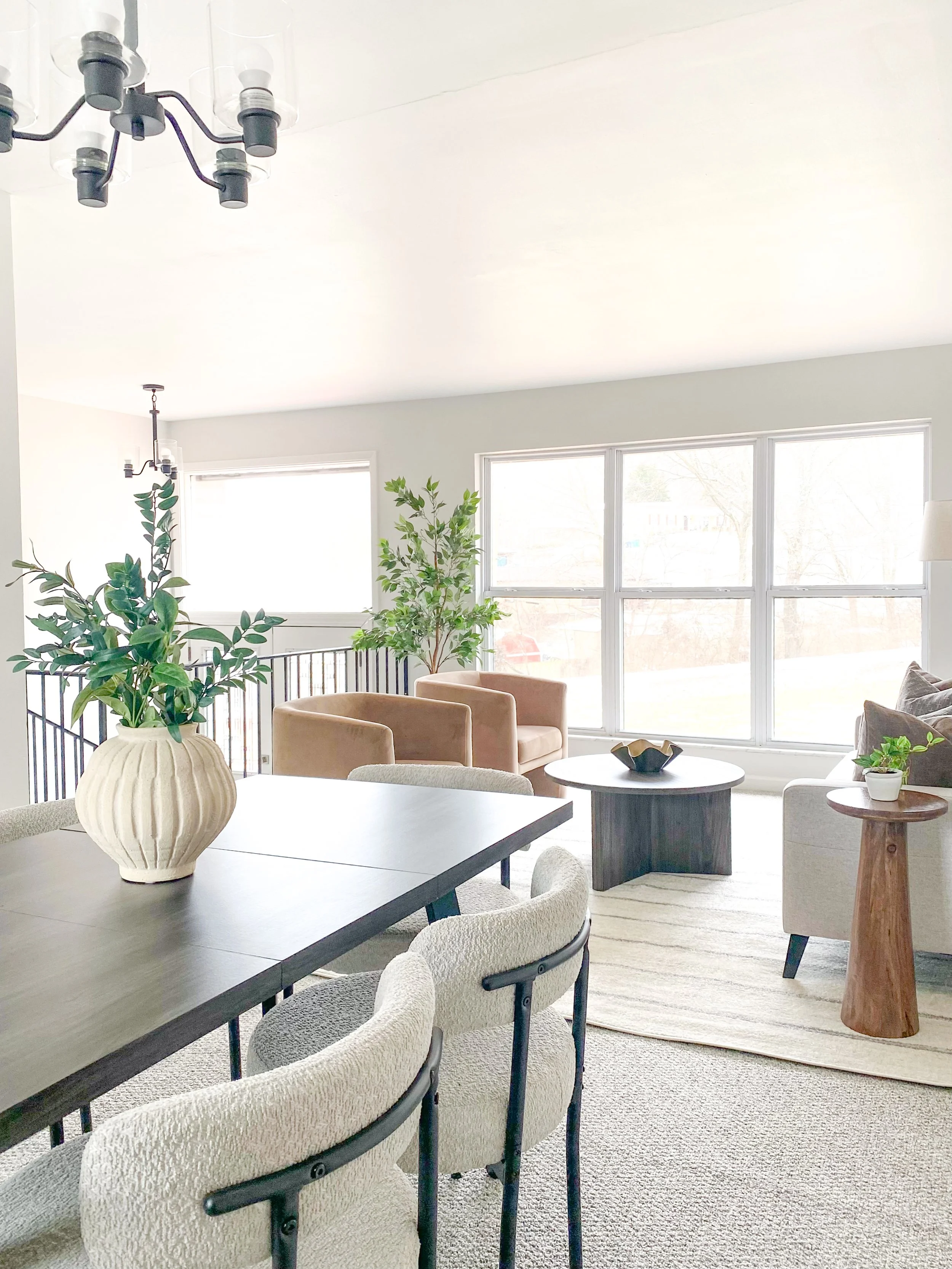 Bright living and dining room with large windows, green plants, dining table with chairs, beige and gray seating, and modern decor.
