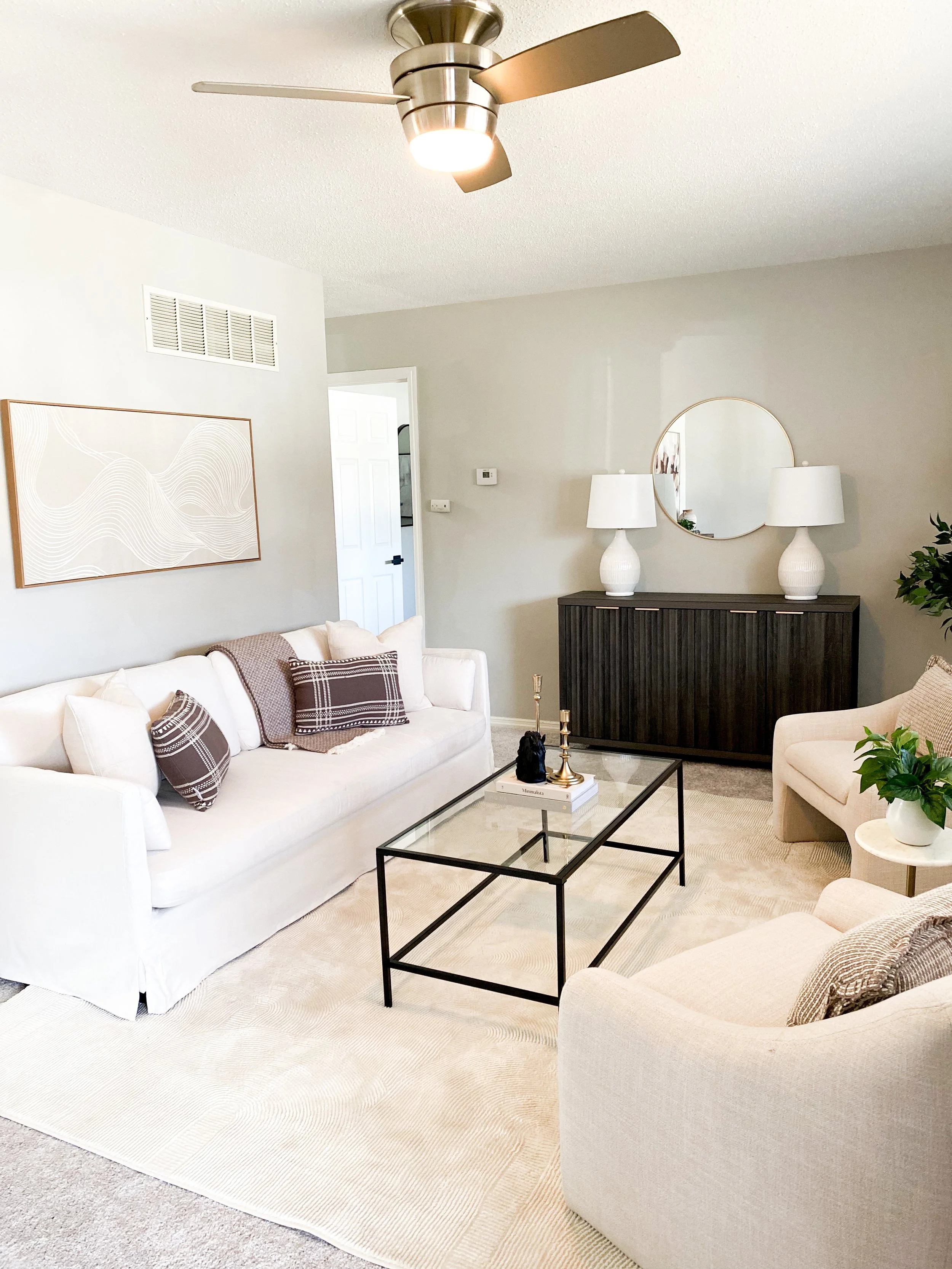 Bright living room with white sofa, beige armchair, glass coffee table, modern white lamps, wall art, round mirror, black sideboard, ceiling fan, and potted plant.