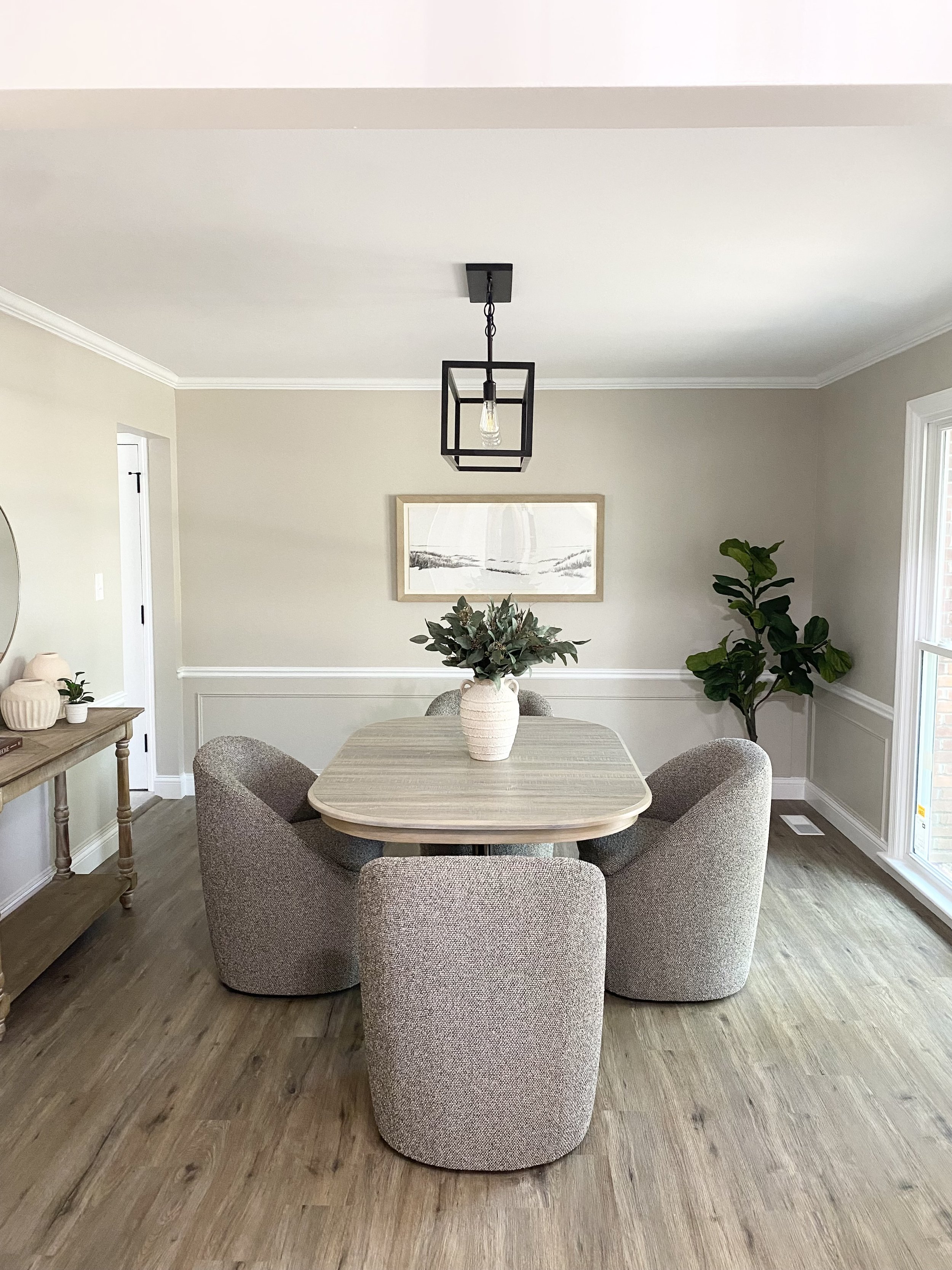 Dining room with beige walls, four upholstered chairs around a rectangular table with a vase of green foliage, modern black geometric light fixture, sideboard with vases on the left, wood flooring, and three windows on the right, natural light illumi