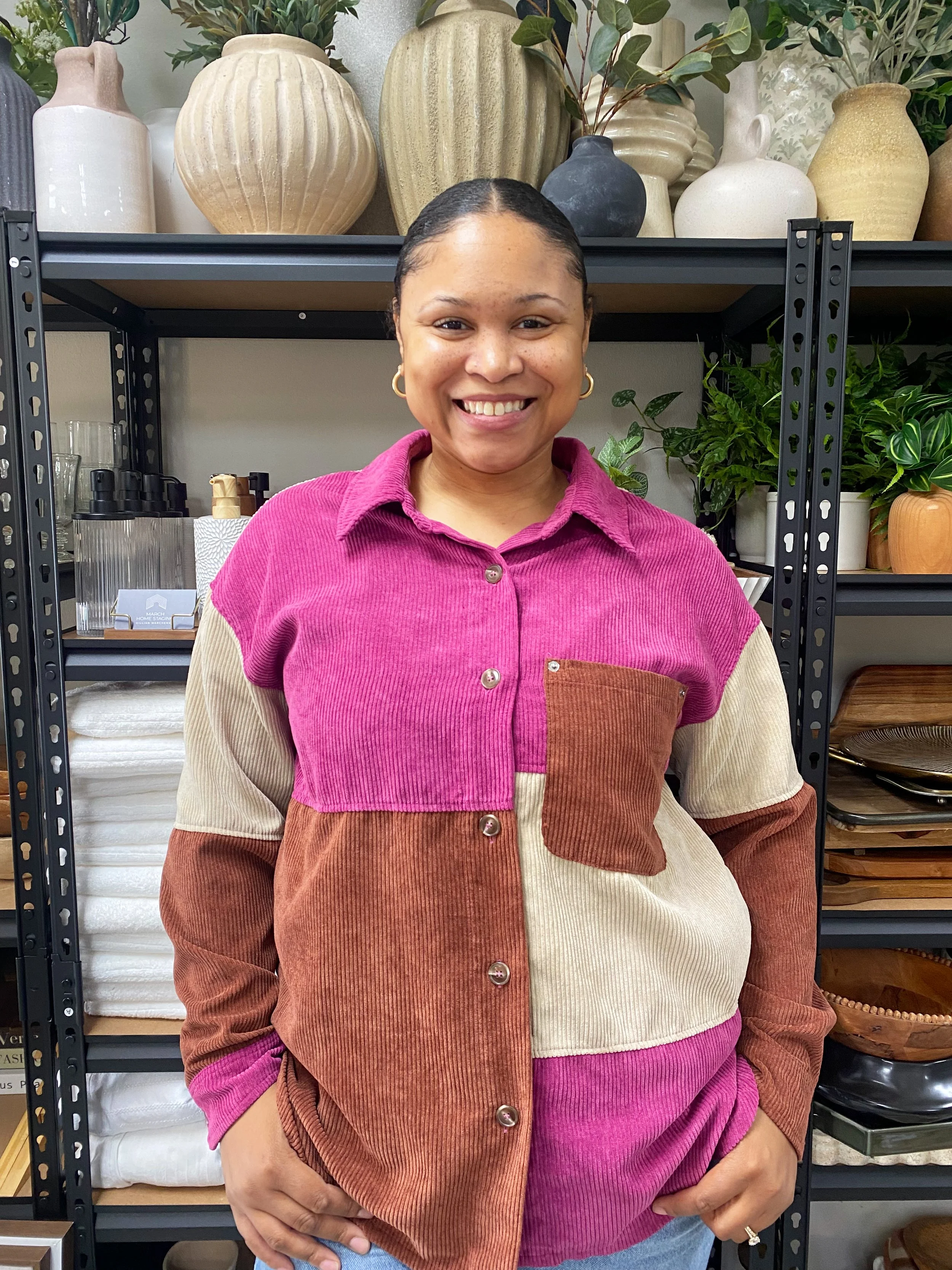 A smiling woman with short hair in a pink, tan, and rust color patchwork corduroy shirt standing in front of shelves with decorative vases and plants.