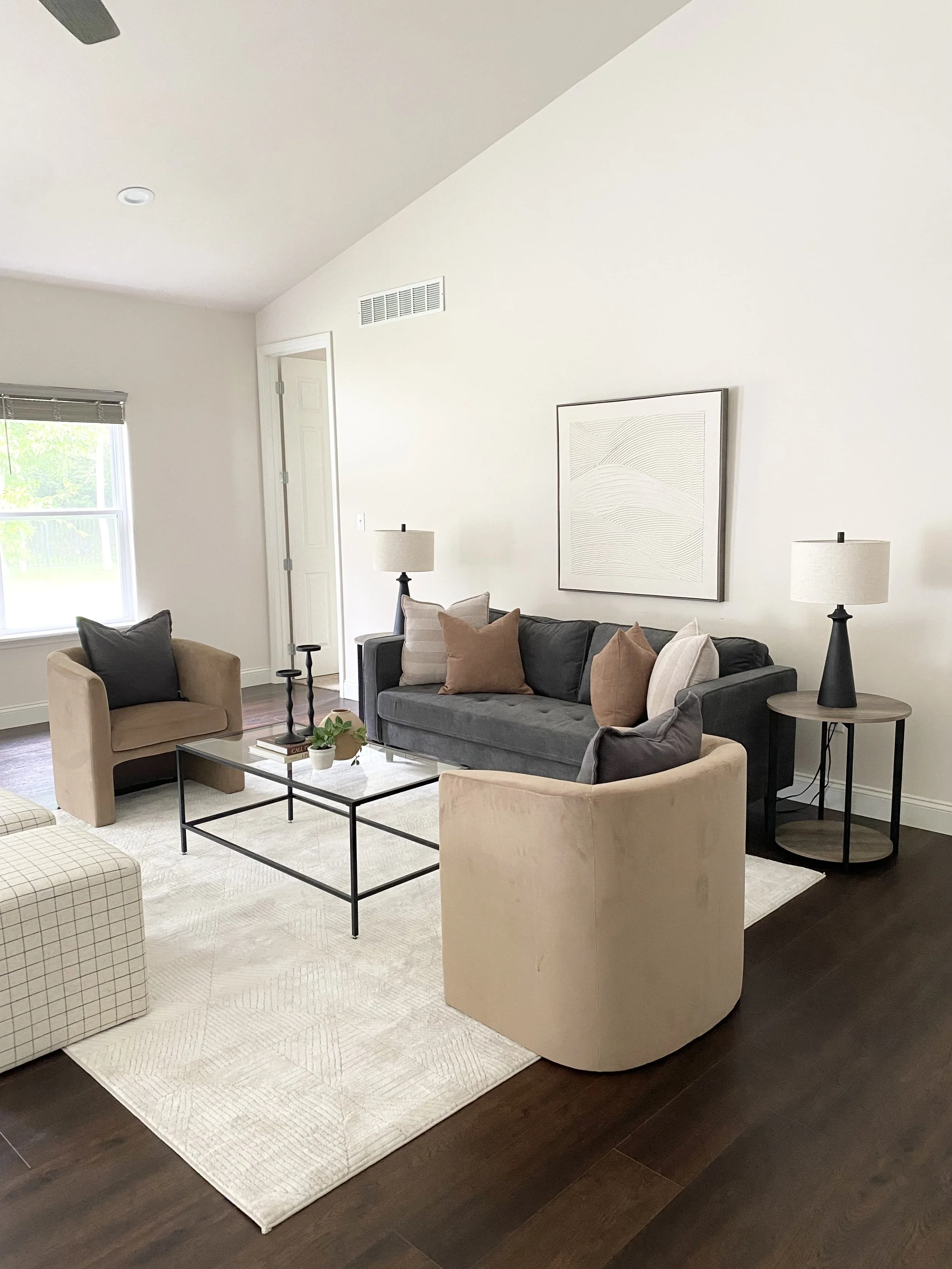 Living room with beige armchairs, a black sofa with cushions, a glass coffee table, and standing lamps, with natural light coming through a window.