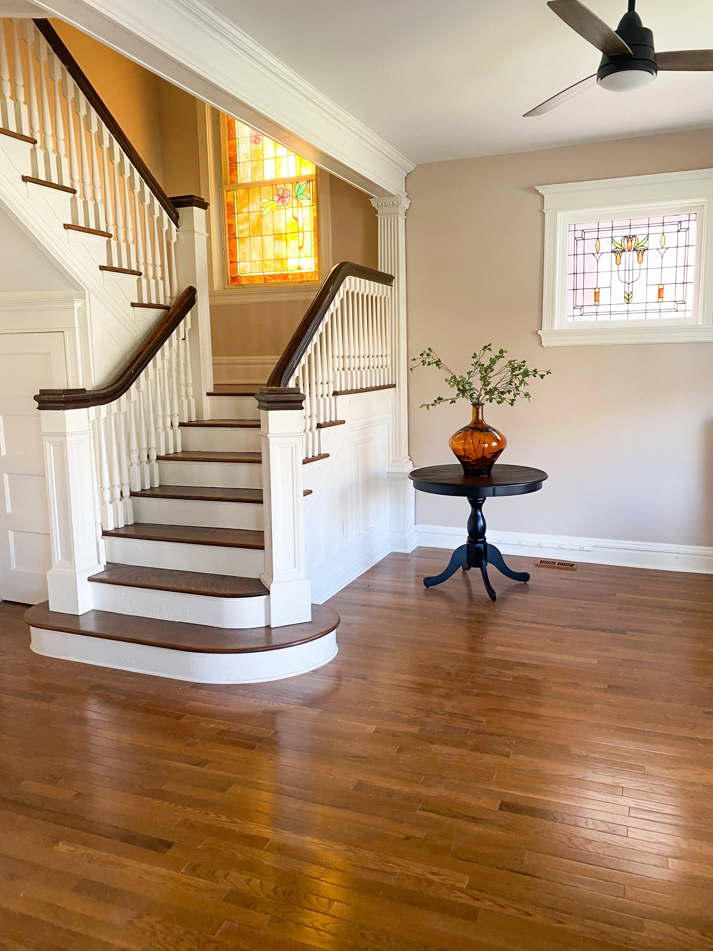 A staircase with white wood paneling and dark wooden handrails, a round black table with a glass vase filled with greenery, and stained glass windows in a home interior with hardwood floors.