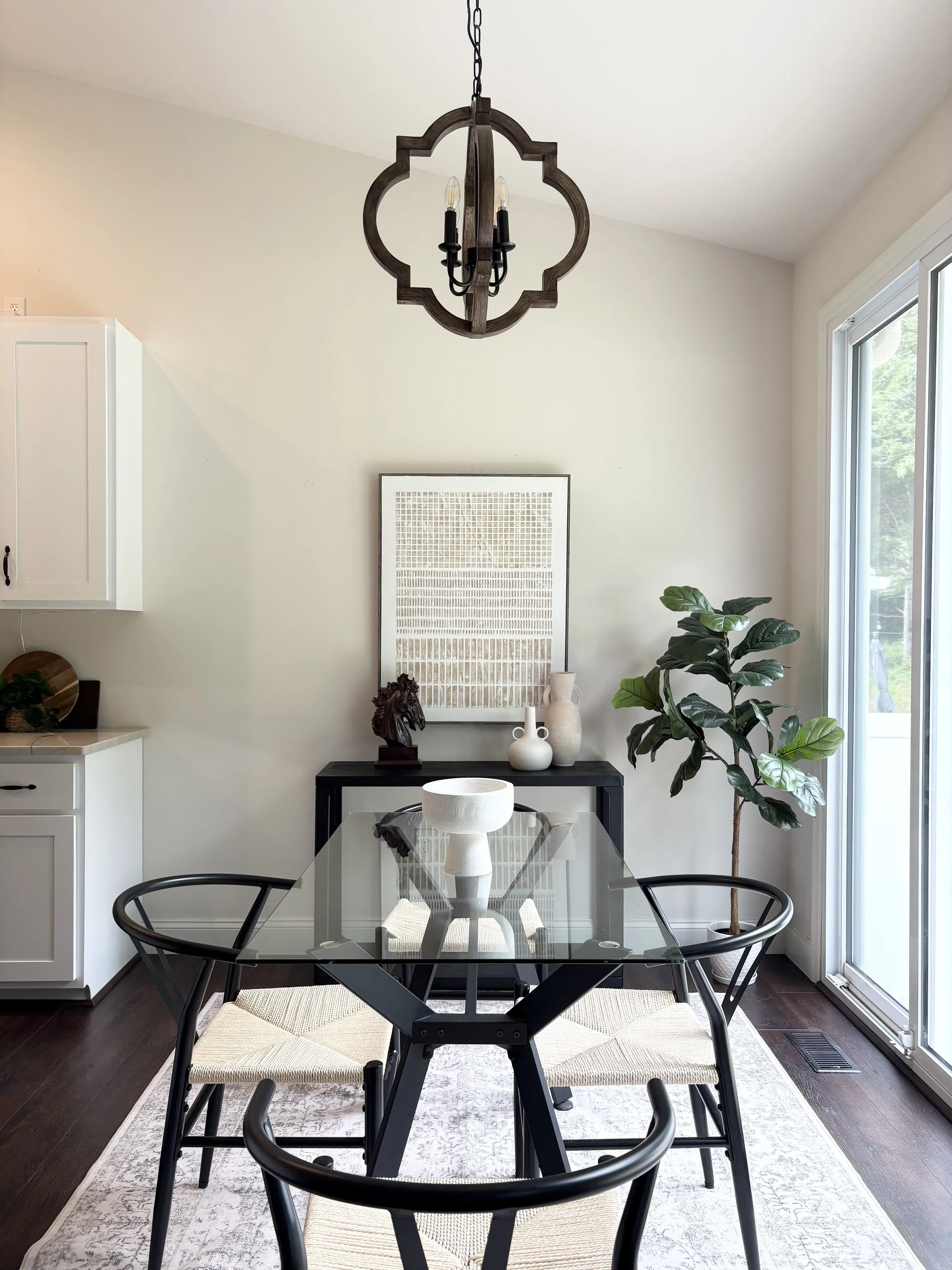 Modern dining area with glass table, four black chairs with woven seats, a black sideboard with decorative vases and sculpture, a large potted plant, a framed artwork, a wooden chandelier, and sliding glass door leading outside.