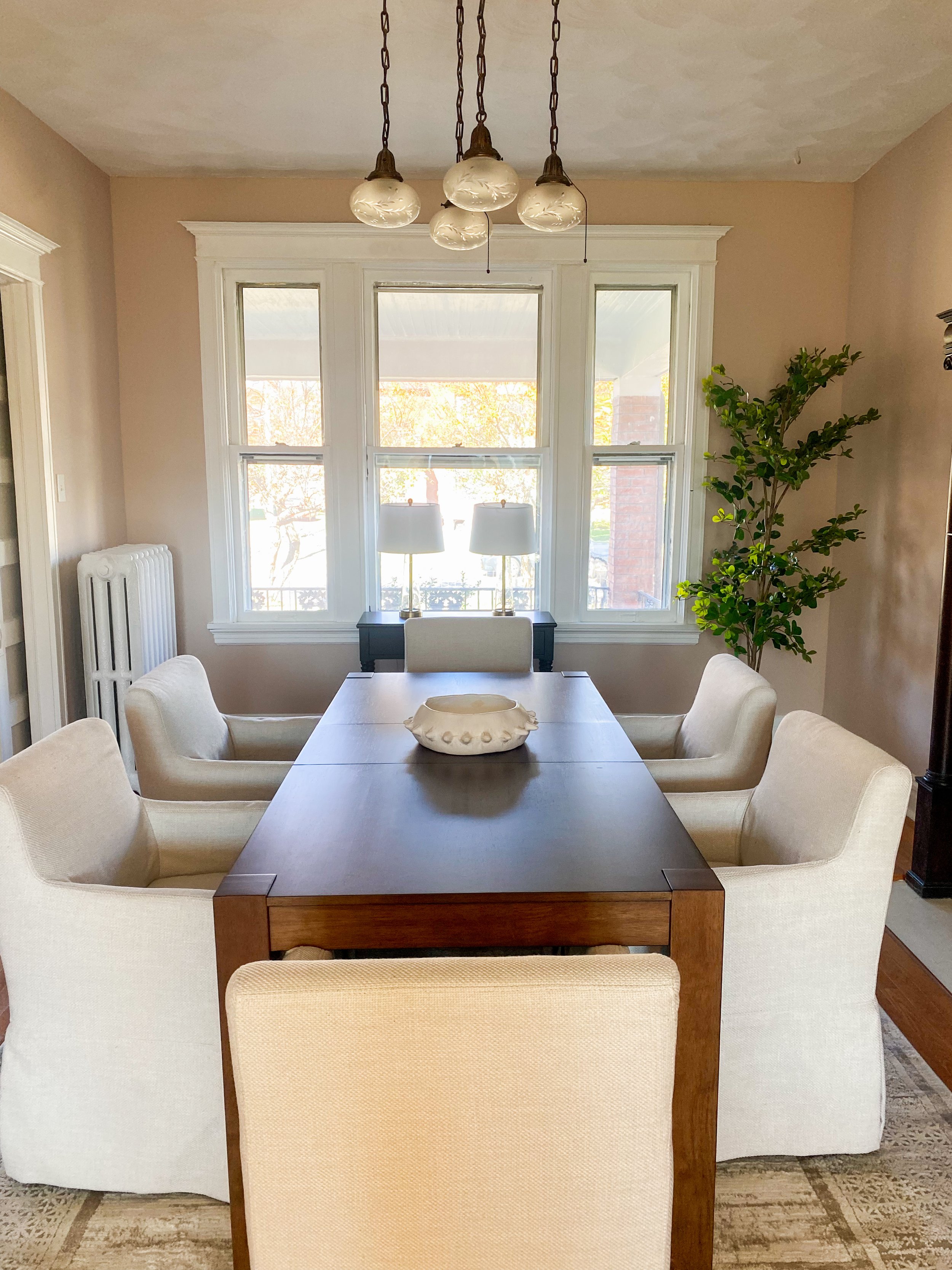 Dining room with a wooden table surrounded by six cream-colored upholstered chairs, a white ceramic bowl on the table, large windows with white trim and two lamps on a black side table, green leafy plant in the corner, and a ceiling light fixture wit