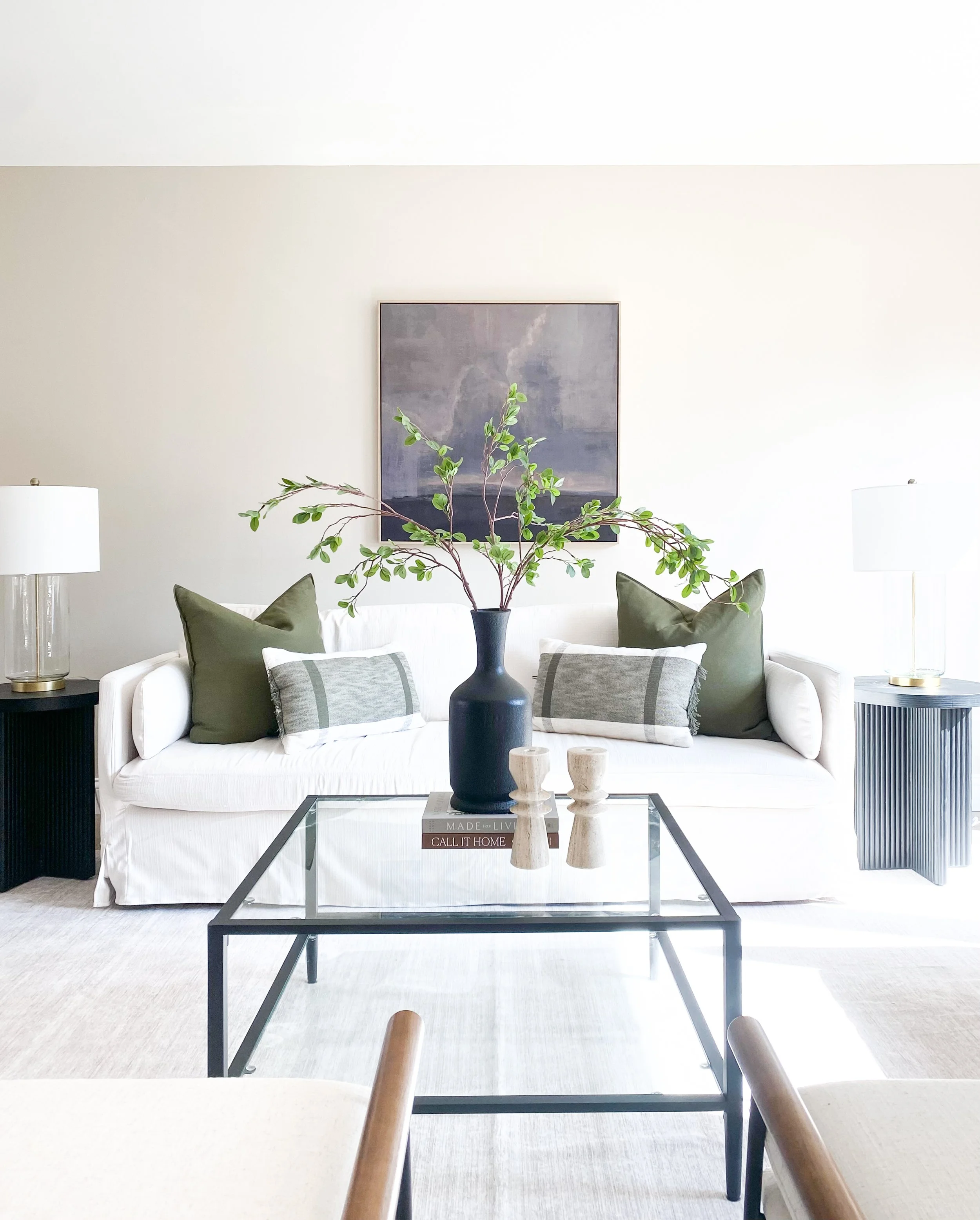 Modern living room with white sofa, green and gray pillows, black coffee table with decorative items, black side tables with lamps, abstract artwork on wall, and large vase with green branches.