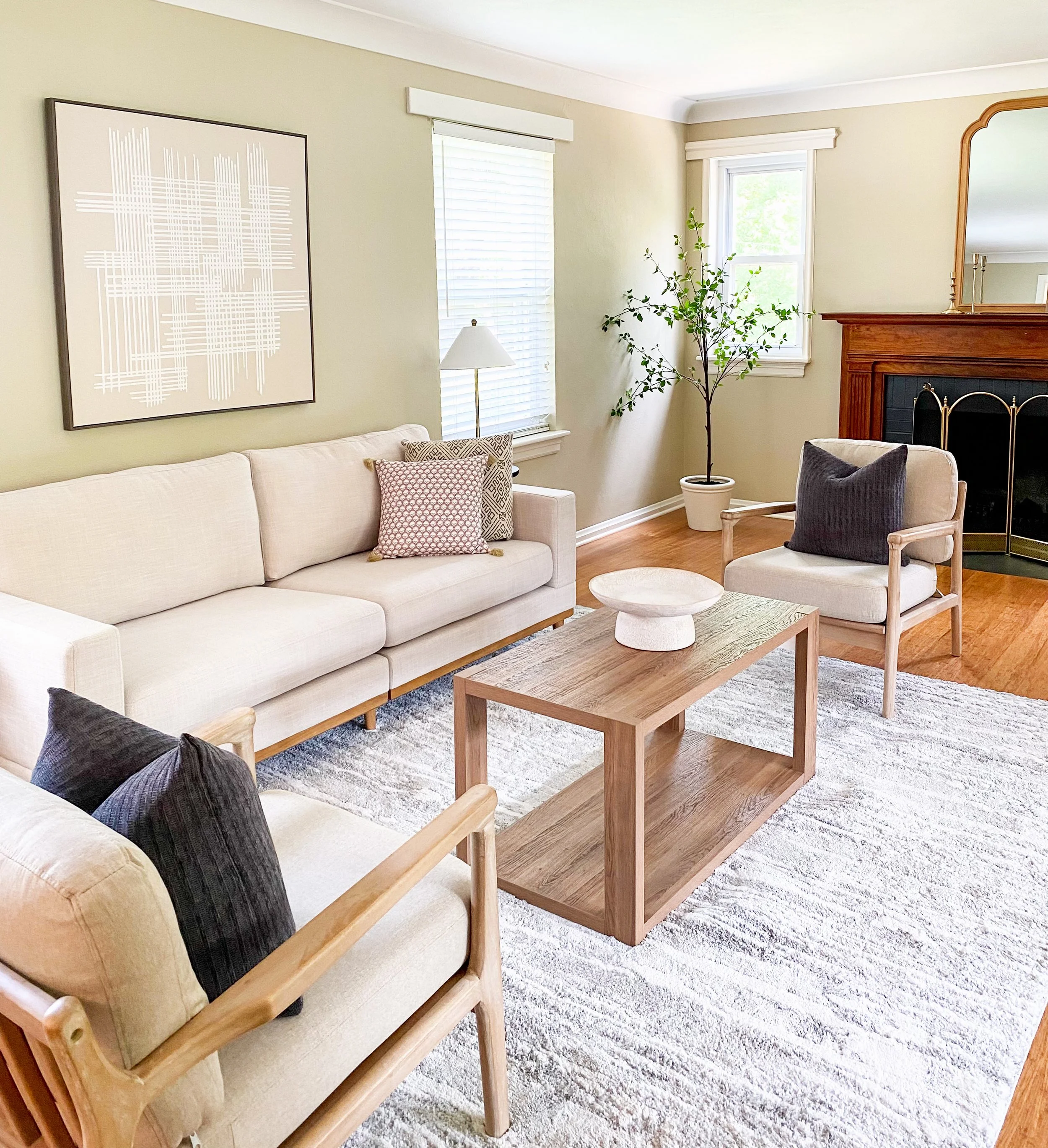 A living room with a beige sofa, two armchairs with black pillows, a wooden coffee table with a decorative white vessel, a white area rug, a potted plant in the corner, a fireplace with a wooden mantel, and large windows with blinds
