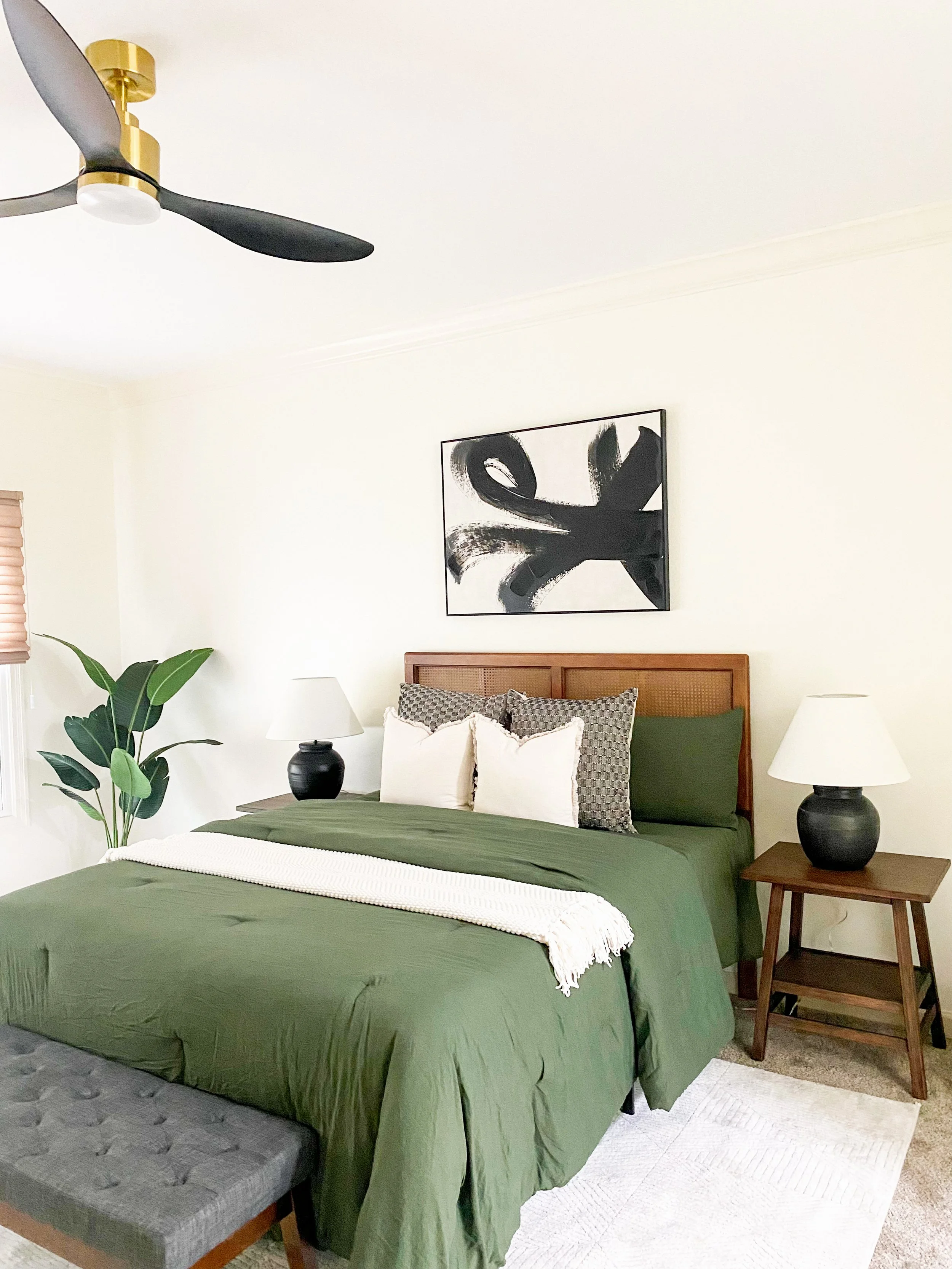 A bedroom featuring a bed with a green bedspread, multiple pillows, and wooden headboard, flanked by two matching bedside tables with black lamps, a potted plant on the left, a modern black and white abstract painting above the bed, a gray bench at t