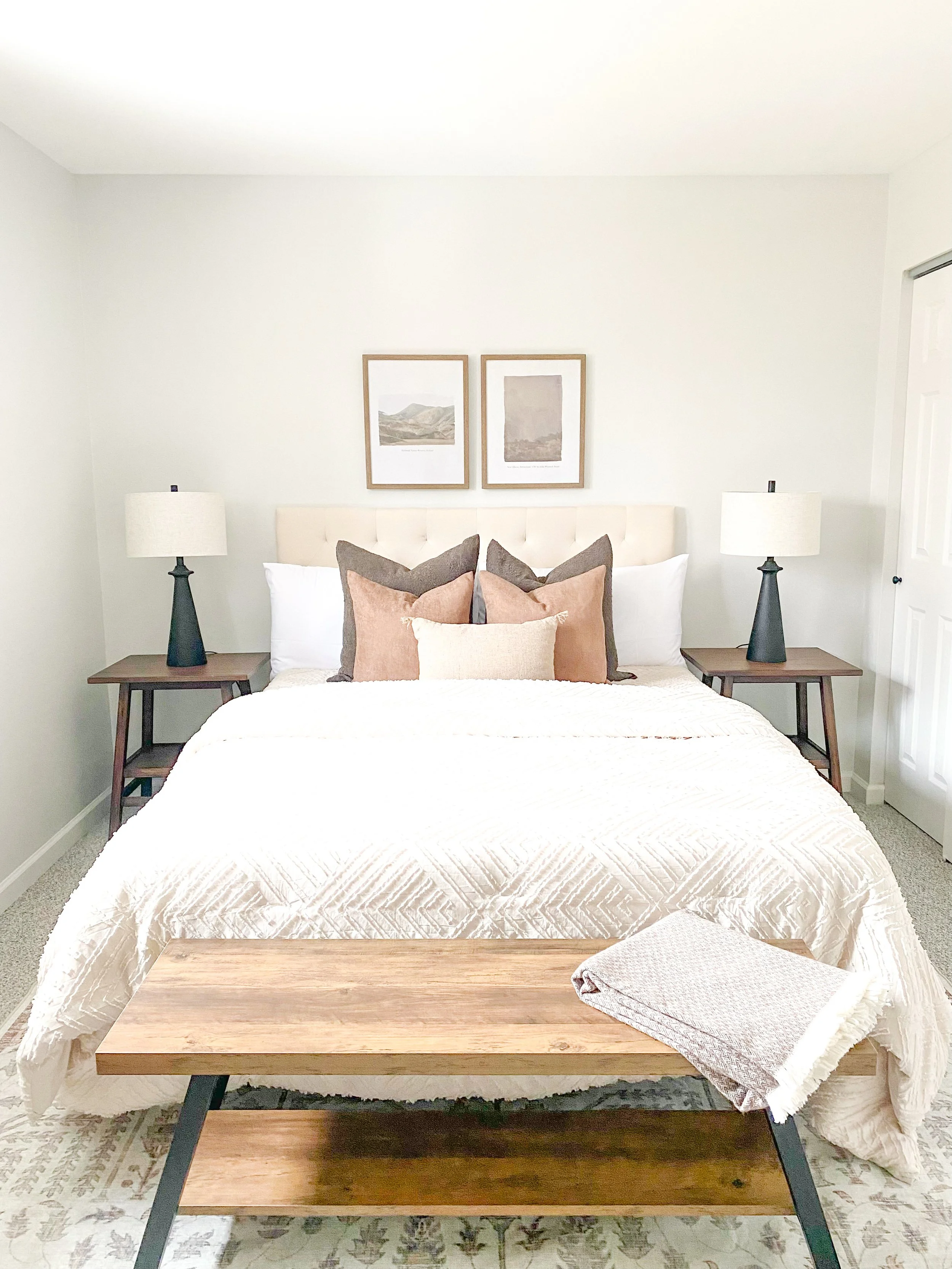 A bedroom with a neatly made bed, two side tables with lamps, framed artwork above the bed, and a blanket folded on a wooden bench at the foot of the bed.