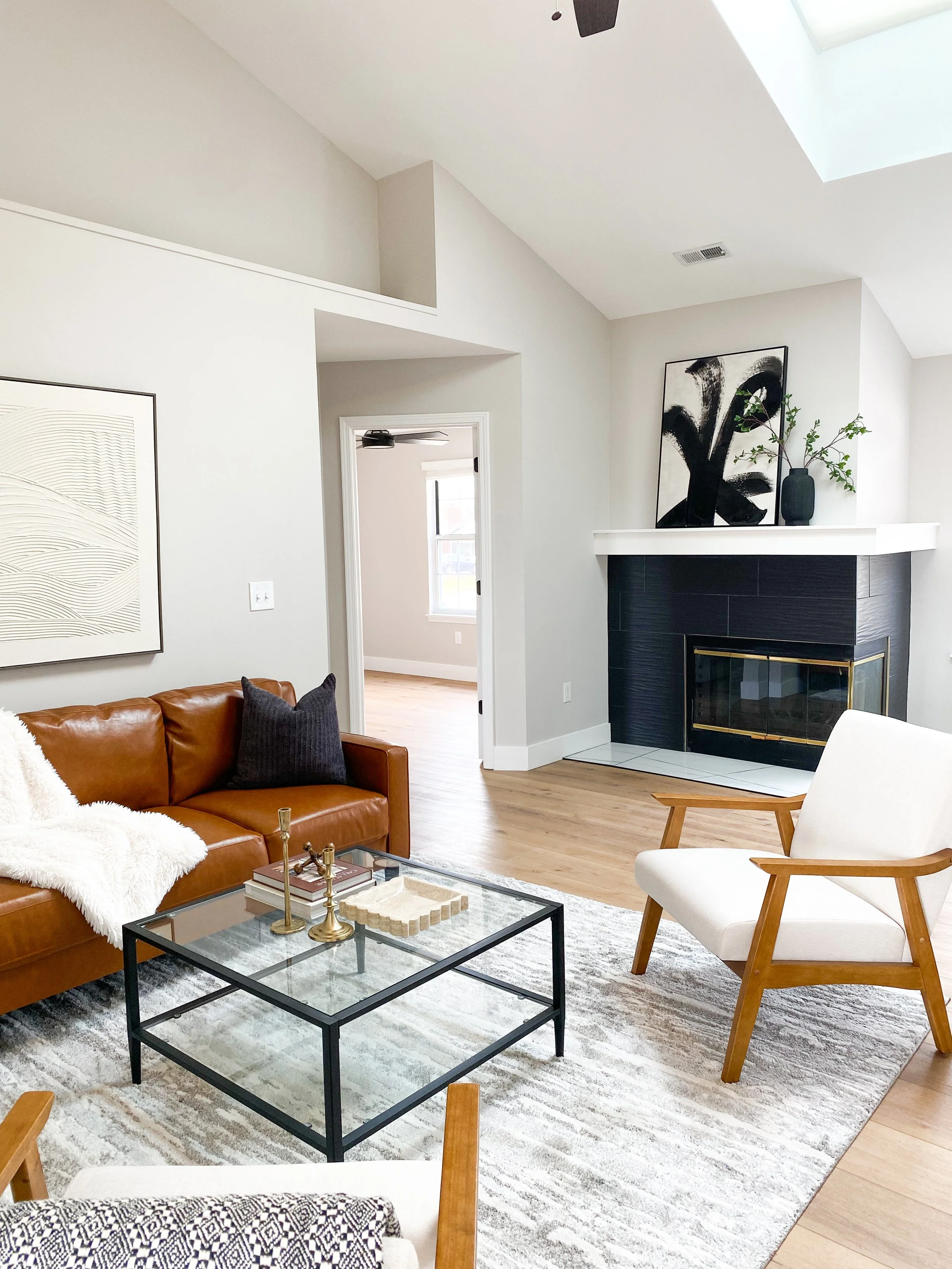 The image shows a modern living room with a brown leather sofa, a white armchair with wooden arms, a glass coffee table with gold accents, a patterned area rug, a fireplace with black tiles, and minimalist artwork and decor, including a black vase wi