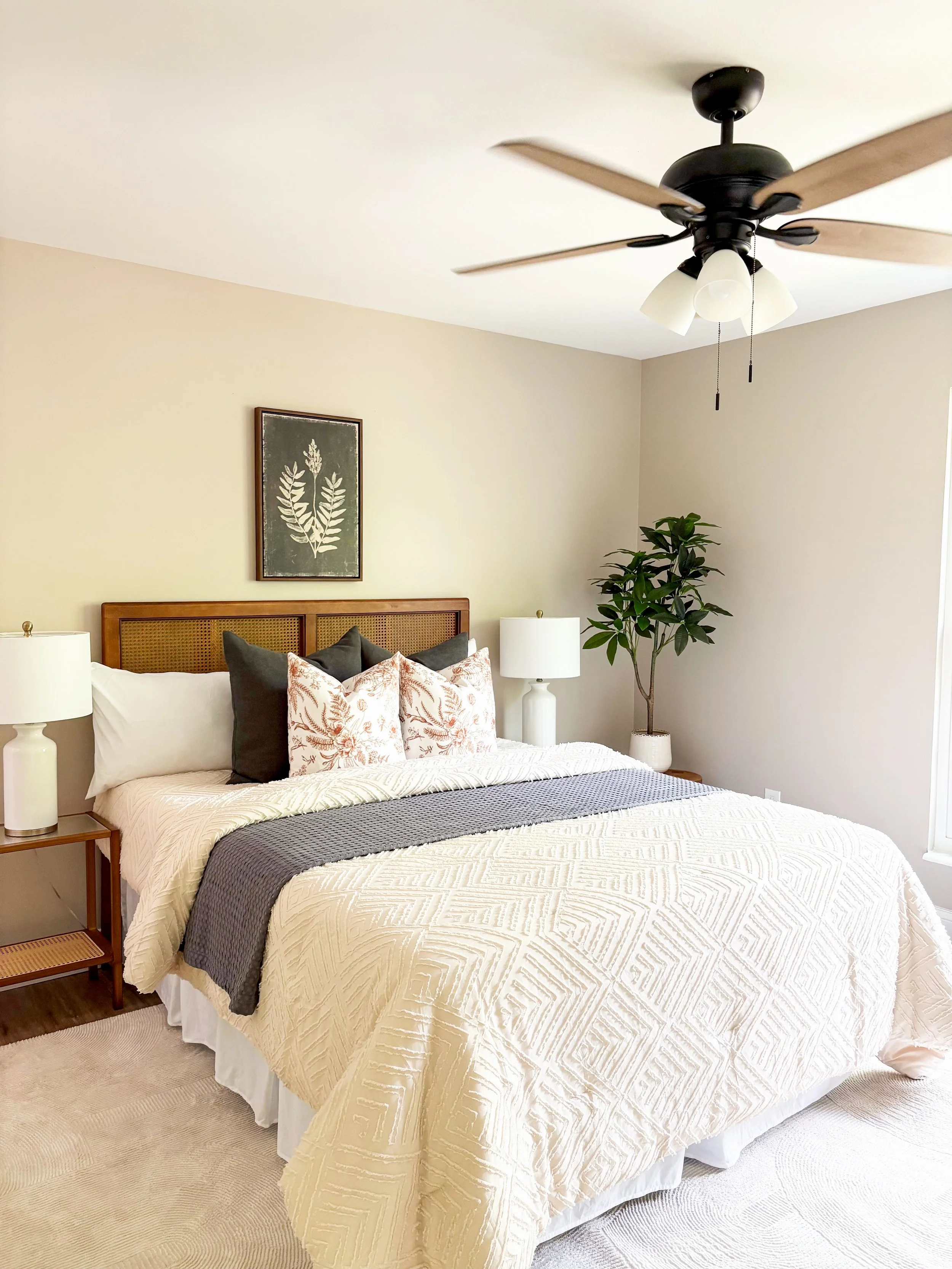 A bedroom with a bed that has a wooden headboard, white bedding, and decorative pillows. There are two white nightstands with lamps, a framed botanical artwork above the bed, a ceiling fan with light fixtures, and a potted plant in the corner.