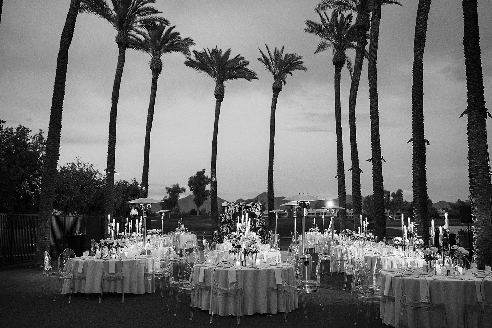 Outdoor wedding reception and event with round tables covered in white tablecloths, decorated with candles, flowers, and glassware, set among tall palm trees at dusk, ghost chairs.